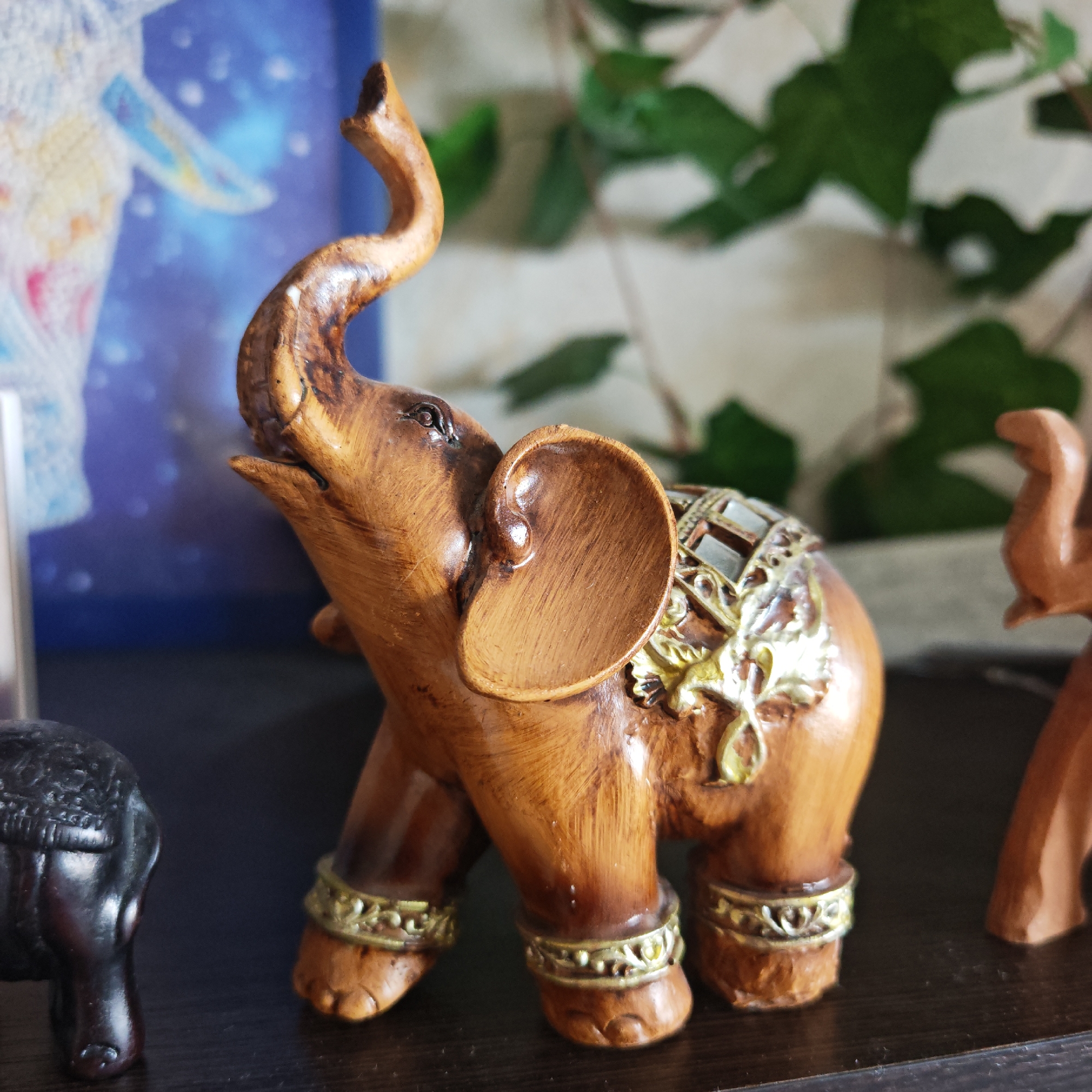 The image depicts a wooden elephant figurine with its trunk raised. It has decorative gold-colored embellishments on its legs and back. The figurine is positioned on a dark surface, possibly a shelf. In the background, there is a blurred image, which appears to be a print or photograph. Green leafy foliage is partially visible behind the elephant. Another smaller, darker elephant figurine is seen partially in the lower left corner. The presence of figurines, particularly elephants, is insufficient to geolocate this image to Rostov, Russia without additional contextual information.