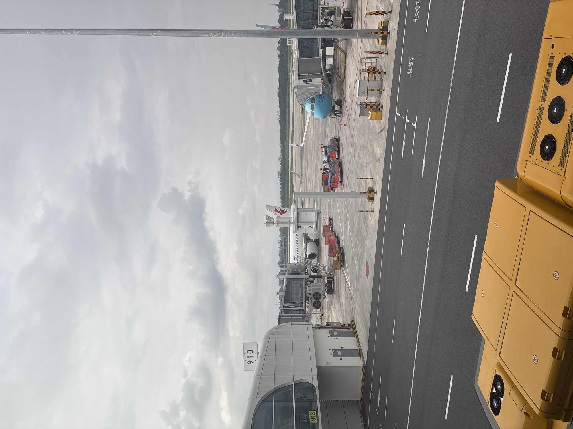 An outdoor, overcast, eye-level view of an airport tarmac at Baiyun District, China shows two airplanes, one white and one blue, connected to jet bridges. The white airplane, with the Eastern Airlines logo, is at gate 913. The blue airplane is at an adjacent gate. The tarmac is marked with white lines and arrows. Various airport vehicles and equipment, including luggage carts and what appear to be baggage tugs, are present. In the foreground, the yellow tops of airport service vehicles are visible. A tall, silver light pole stands on the right side of the frame. The sky is cloudy and grey.