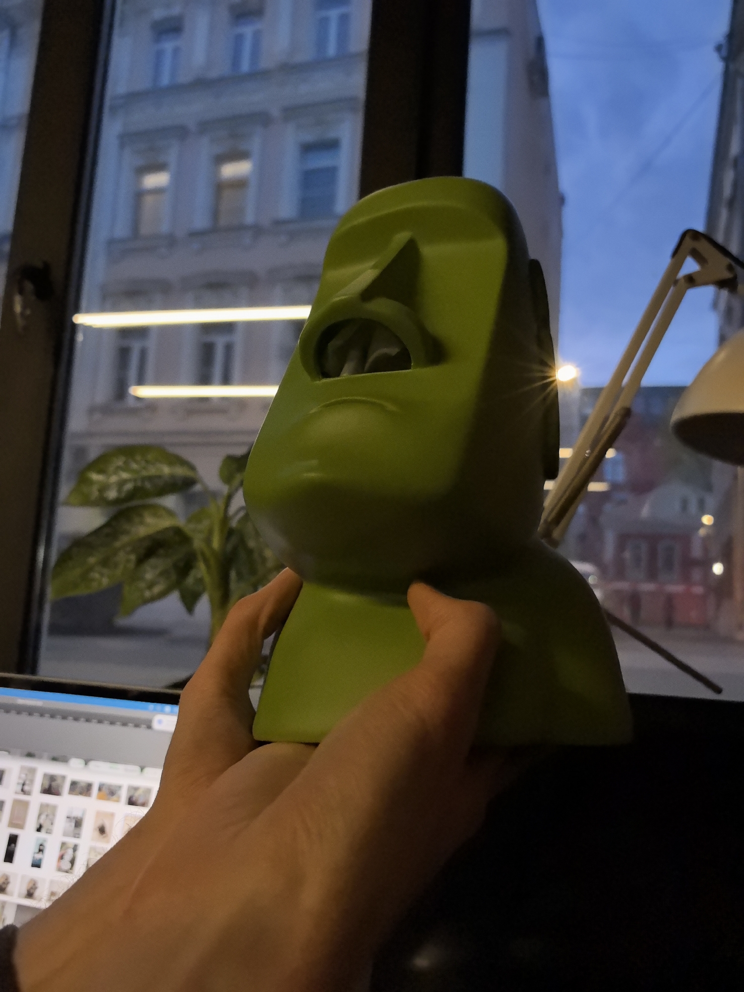 A person is holding a green Moai head tissue box in front of a window. Through the window, buildings are visible, including a multi-story building with illuminated windows across the street. There's a desk lamp with a white lampshade to the right of the Moai head. A computer monitor is partially visible in the lower left corner of the image, displaying a screen with numerous images or thumbnails. A small green plant is present on the desk near the window. Based on the architectural style and general urban layout, the location is possibly Moscow, Russia.
