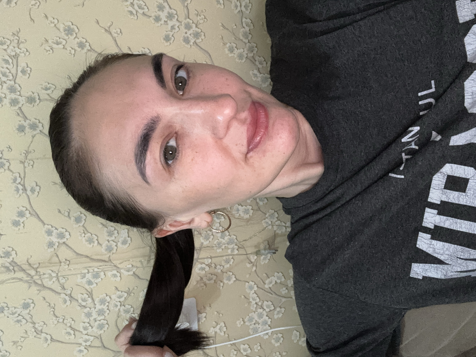A woman is posing for a selfie indoors. She has dark hair pulled back in a ponytail, light skin, and is wearing a gray t-shirt with white lettering. She is smiling slightly. She has hoop earrings in her ears. The background is a light beige wall with a floral pattern. The location is not possible to determine based on the image.