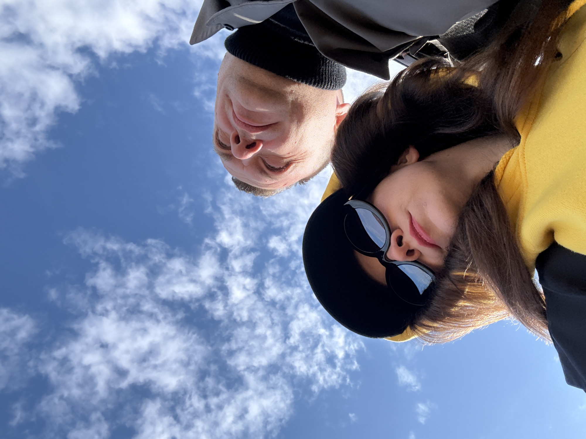 An extreme low-angle photograph captures two individuals, a man and a woman, looking directly down towards the camera, set against a vibrant blue sky dotted with scattered white clouds. The man occupies the upper left portion of the frame, wearing a dark jacket and a black knitted beanie. His face is illuminated by bright daylight, showing a slight, gentle smile and squinted eyes, possibly due to the sun. To his right, the woman wears a bright yellow garment, possibly a hoodie, along with a dark hat and large, round black sunglasses that obscure her eyes. Her dark hair cascades around her face and shoulders. Both subjects are positioned as if leaning over the camera, creating a worm's-eye view. The clear sky and strong illumination indicate it is daytime with fair, sunny weather. The general atmosphere suggests an outdoor setting in Chaikovsky, Russia. No text is visible in the image.