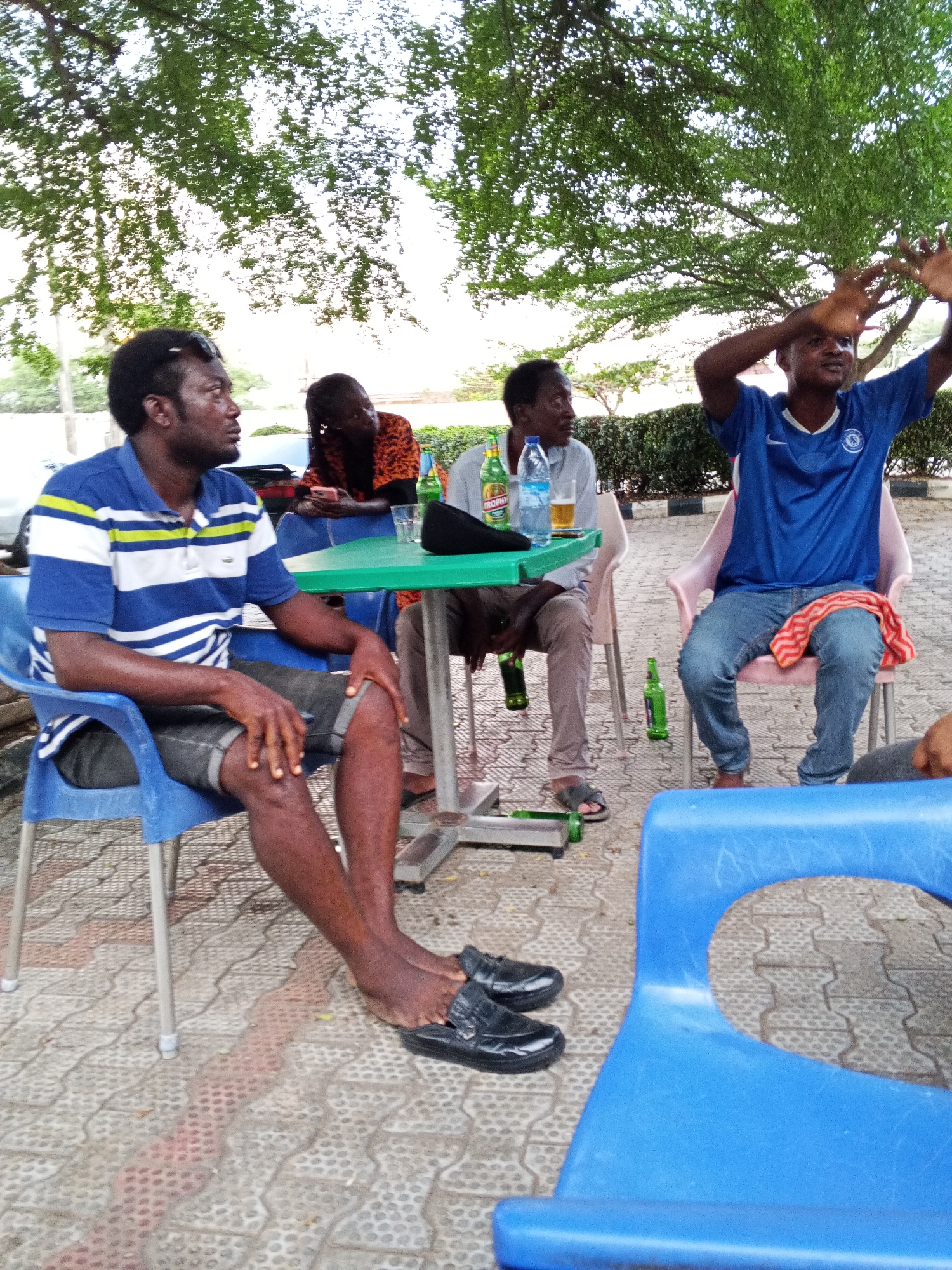 Four men are seated at a light green table under the shade of a tree in Lokoja, Nigeria. The man in the foreground wears a blue and white striped polo shirt, black shorts, and black shoes, seated in a blue plastic chair. To his left are three men; one is looking at his phone. The man to the right of the man on the phone is wearing a gray shirt with a beer in front of him. The fourth man, furthest right, is gesturing with both hands in the air. There are bottles of beer and water on the table. Empty green beer bottles lay on the ground. The ground is covered in a patterned light gray paving stone.
