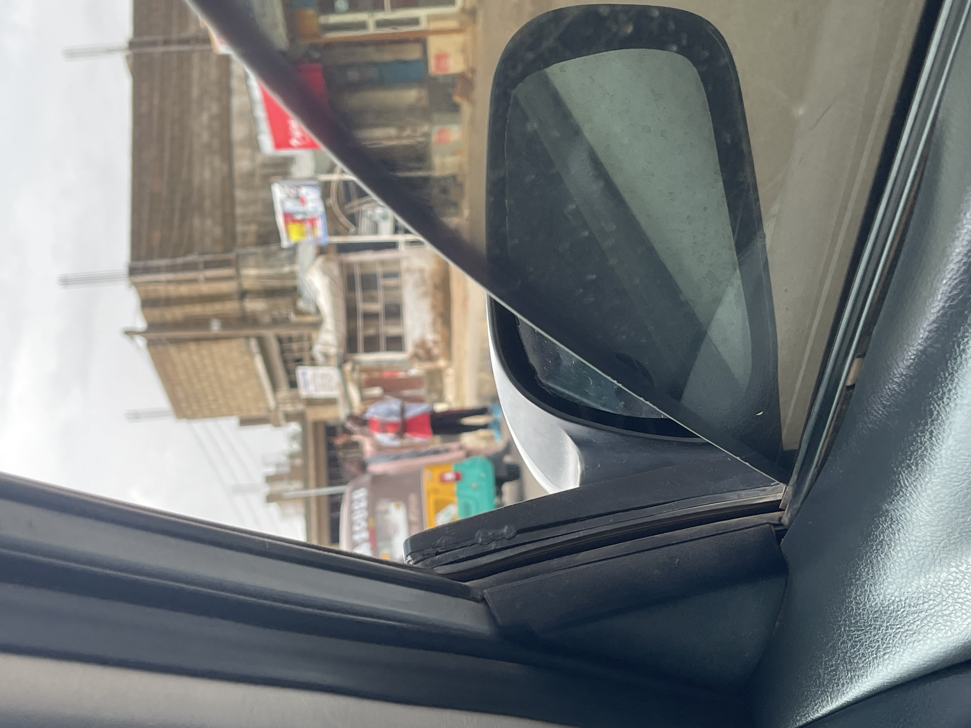 View from inside a car, looking out at a street scene. The car's side mirror is in the foreground. Outside, there are multi-story buildings with shops and signs. People are walking on the sidewalk. The location appears to be in Jos, Nigeria.