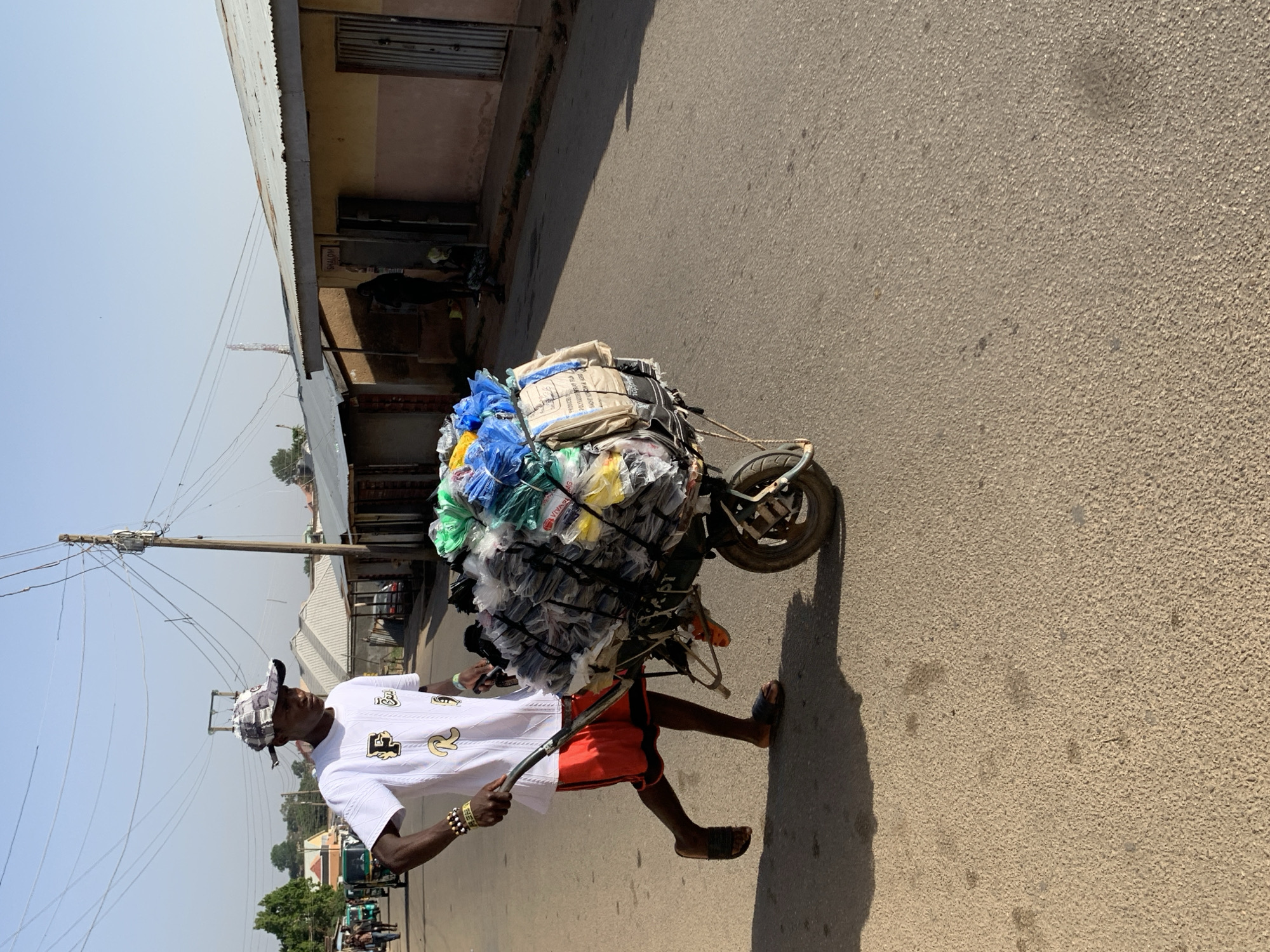 A man in a white shirt, red shorts, and sandals pulls a heavily laden one-wheeled cart along a paved road in Jos, Nigeria. The cart is piled high with various items wrapped in plastic bags and tied with rope. The man wears a patterned hat and bracelets. The background includes buildings, power lines, and trees under a clear sky.