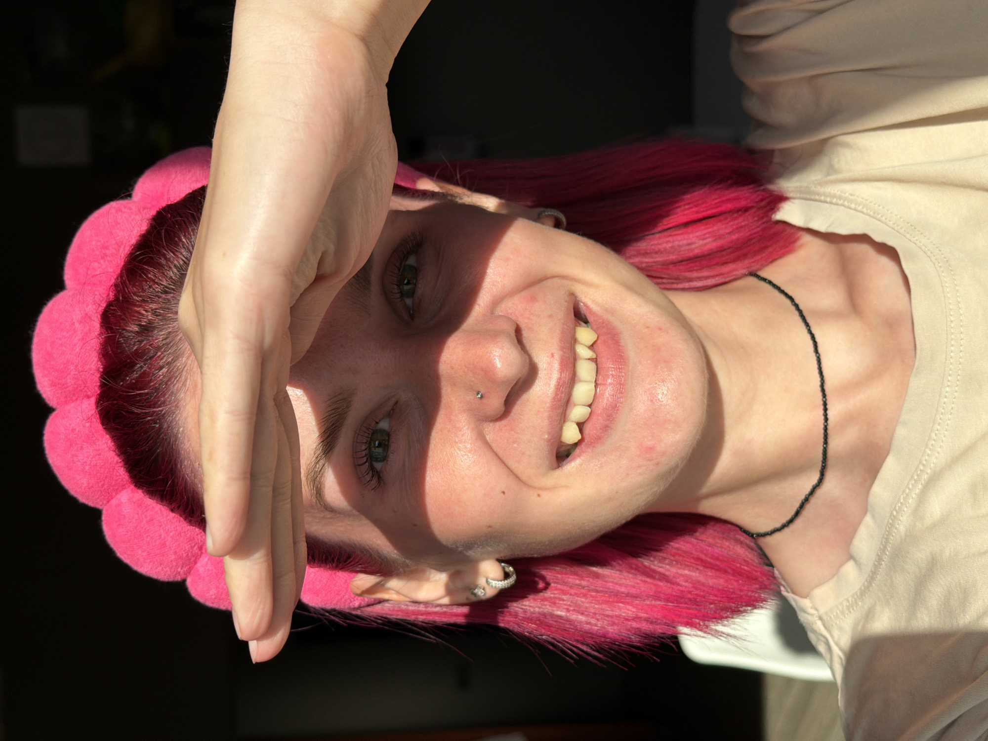 A young woman with bright pink hair is shown in a close-up portrait, with her head tilted and one hand covering her forehead. Sunlight streams across her face, creating strong shadows. She is wearing a fuzzy, bright pink headband with scalloped edges. Her expression is a mix of a smile and a squint against the light, with her mouth open to reveal teeth. A small silver nose stud is visible. She has on a light-colored t-shirt and a thin black beaded necklace. The background is dark, suggesting an indoor setting with a strong light source from the side. The woman appears to be in a casual and playful mood. The time of day is likely daytime due to the strong sunlight.