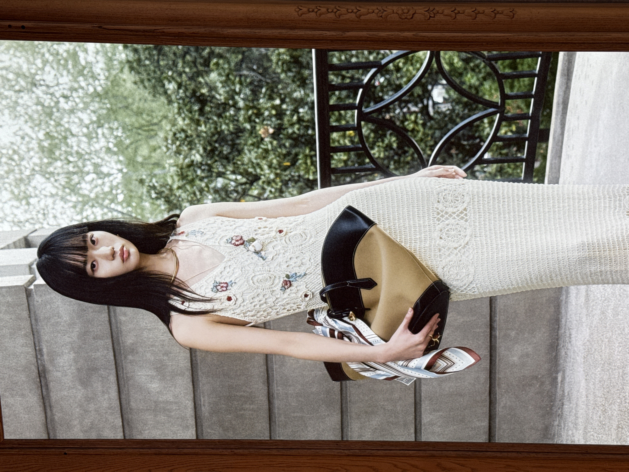 A young Asian woman with straight black bangs and long, straight black hair stands in what appears to be a courtyard or balcony area, framed by a wooden doorway on the right side of the image. She is wearing a sleeveless, cream-colored crocheted dress with floral embroidery. Around her neck, she wears a thin gold necklace. She is holding a large, tan and black tote bag with leather handles, and a patterned silk scarf is draped over one of the bag's straps. The background consists of a textured gray wall, lush green trees and foliage, and a decorative black metal railing. The ground is a light-colored, textured surface, possibly concrete. The overall setting suggests an outdoor, possibly urban environment.