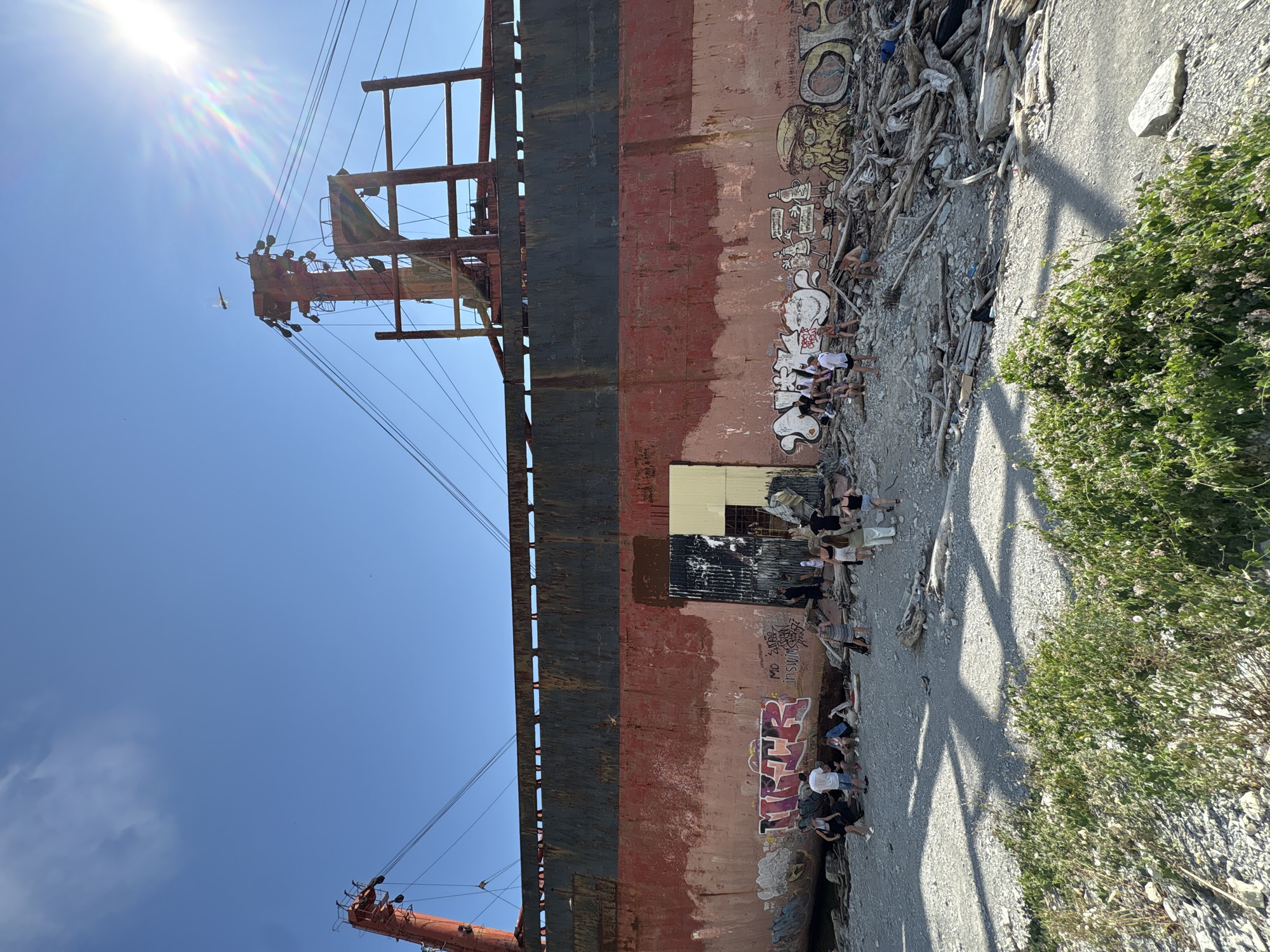 A group of people are gathered on a rocky shore in front of a large, rusted ship that has run aground. The ship has graffiti on its hull. The sky is clear and blue with a bright sun. The scene is located in Kabardinka, Russia.