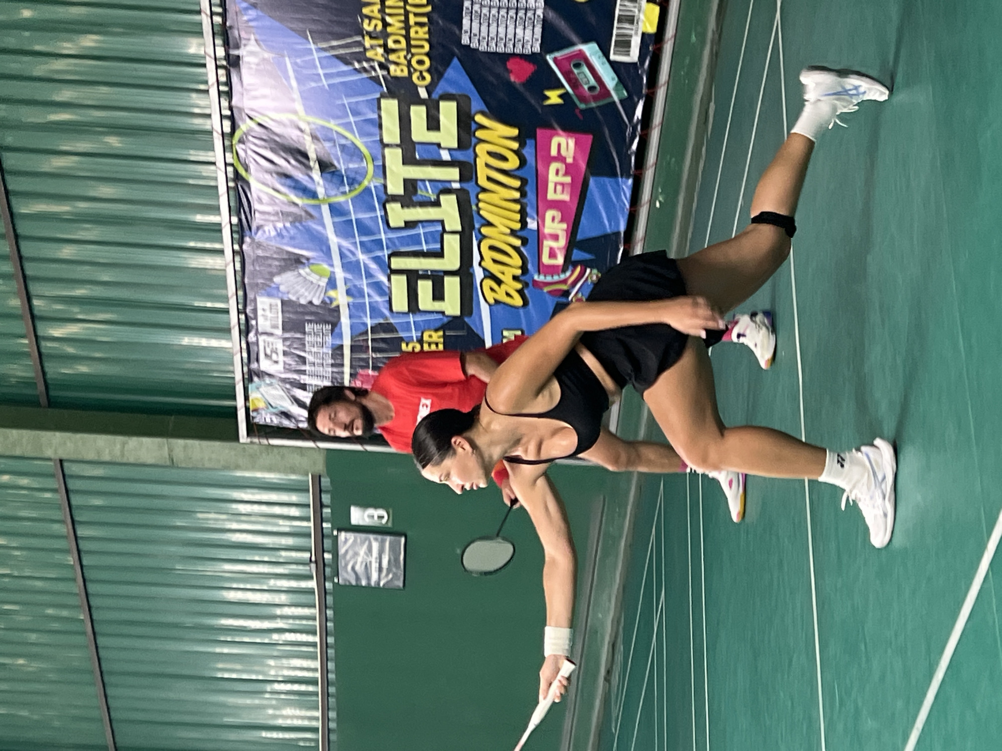 The image shows two people on a badminton court in Ban Bang Rak, Thailand. A woman in the foreground is wearing a black sports bra, black shorts, white socks, and white sneakers. She is in a lunging position, holding a badminton racket. A man in the background is wearing a red t-shirt and dark pants. He is standing behind the woman, looking forward. A banner with "ELITE BADMINTON CUP FP.2" is visible behind them. The court is green with white lines. The background consists of a green corrugated wall.