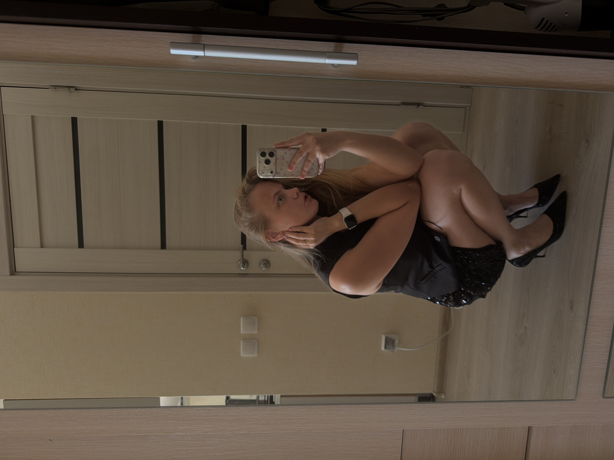 A mirrored reflection shows a woman seated, facing left, taking a selfie with her phone. She has blonde hair and is wearing a black top or dress and black high heels. Her legs are tucked up close to her body. The reflection shows a light-colored door to the left, and light-colored walls with electrical outlets and light switches. The mirror is part of a light-colored wooden structure. There is no indication of the city of Izhevsk, Russia.