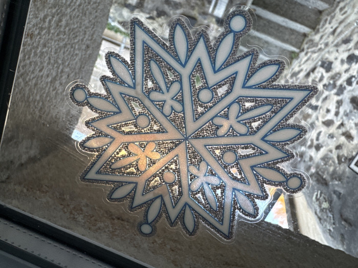 A decorative snowflake decal is affixed to the interior surface of a clear glass window pane. The snowflake design is multi-pointed, featuring white segments outlined in blue, with interior areas filled with textured silver glitter that reflects ambient light, displaying warm gold and orange tones. The decal is oriented slightly diagonally within the frame.
Through the window, a view of an outdoor urban environment in Le Puy-en-Velay, France, is visible. To the left and bottom-left, a robust stone wall constructed from dark, irregularly shaped stones is prominent, leading down to a visible section of stone steps. To the right and top-right, another rough-textured, lighter-colored stone facade is present. The dark frame of the window unit is visible along the top and right edges of the image. The scene is illuminated by daylight, with reflections evident on the glass. No people or active interactions are depicted.