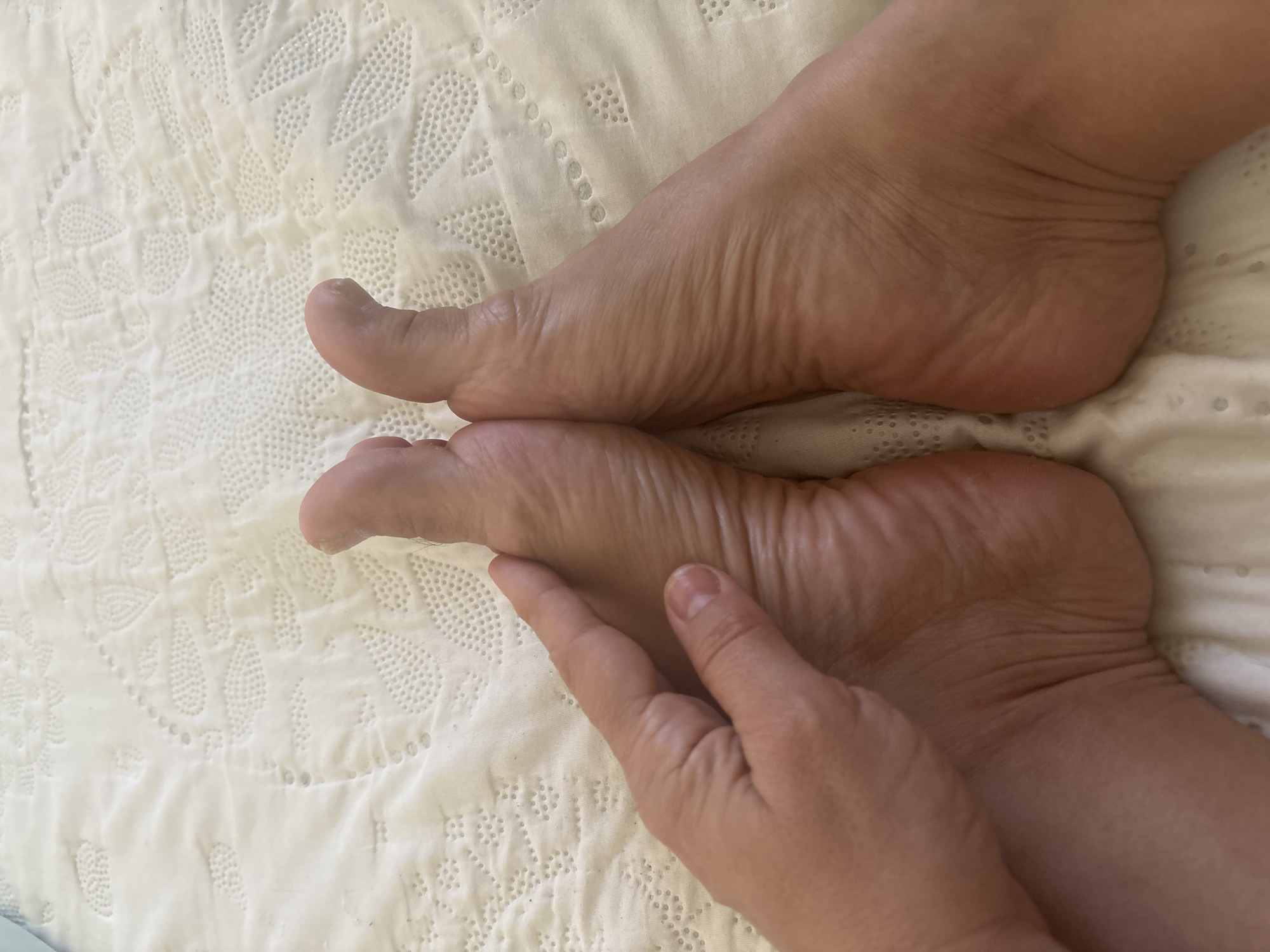 This is a close-up, overhead shot of a pair of bare feet resting on a white, textured surface, possibly a bedspread or blanket. A person's hand is visible on the left side of the image, gently touching the sole of one of the feet. The feet are light-skinned with visible wrinkles on the soles and toes, suggesting they are relaxed. The background has a repeating pattern of embroidered or quilted designs. There is no indication of the city of Lod, Israel, or any other specific location in this image.