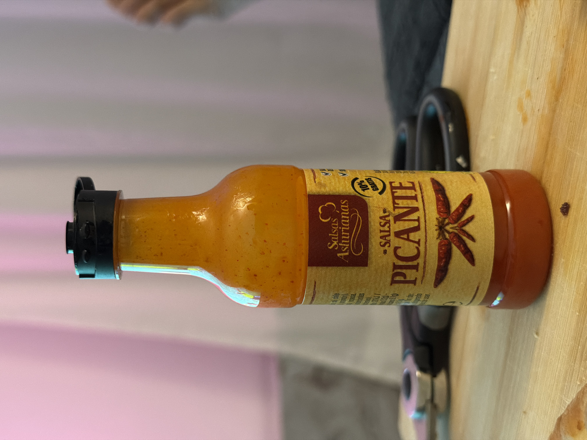 A close-up, slightly elevated shot shows a bottle of orange-red hot sauce, labeled "Salsas Asturianas PICANTE," placed on a light-colored wooden surface. The bottle has a black cap and a yellow label with red text and an illustration of chili peppers. To the left of the bottle, a pair of scissors with black handles and metallic blades is visible, lying on the same wooden surface. The background is softly blurred, with vertical lines of pink and gray fabric creating a gentle drape effect. The overall lighting is soft, with some highlights reflecting on the glass bottle. The scene appears to be indoors, possibly in a kitchen or dining area.
