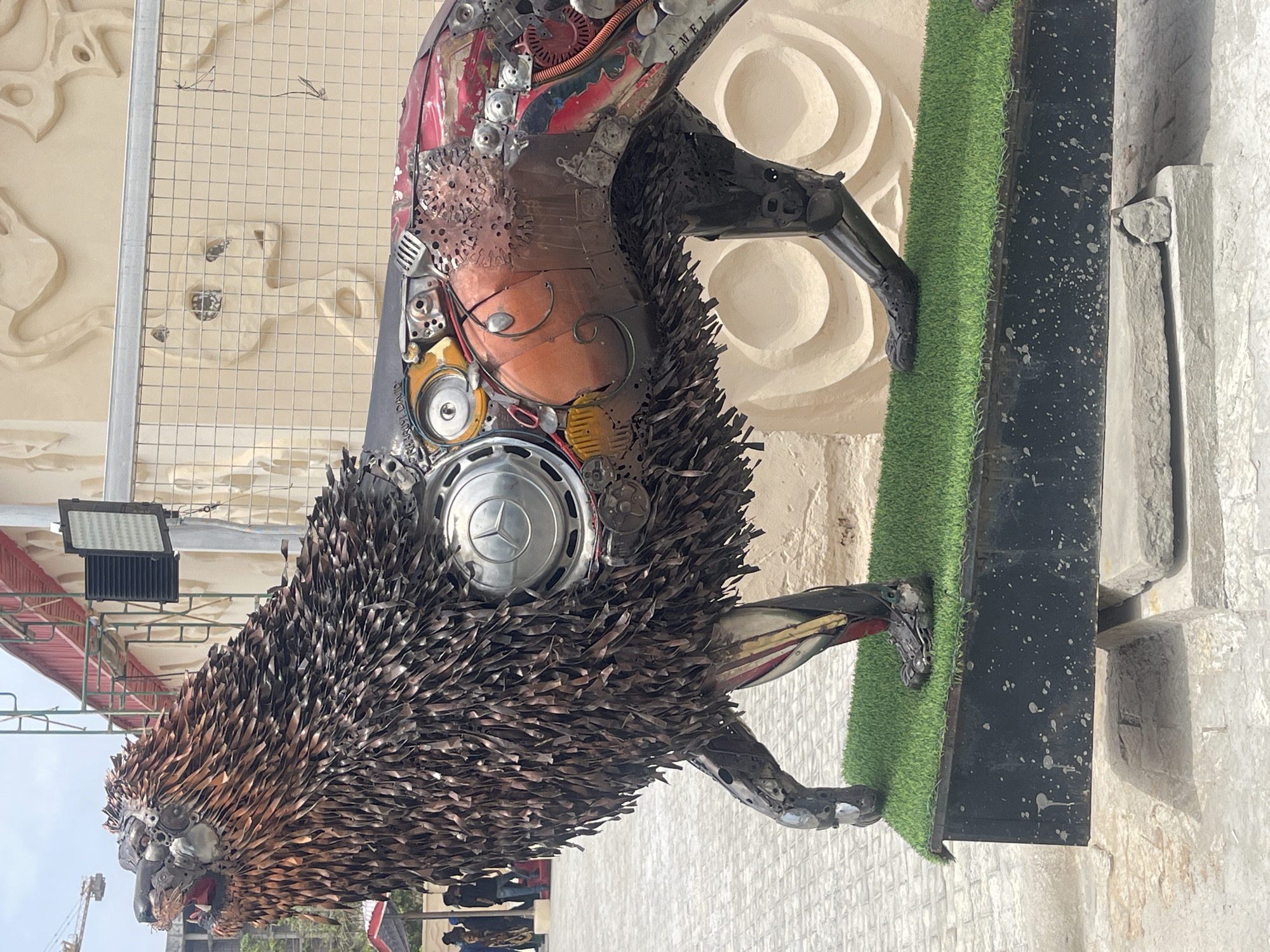 The image shows a metal sculpture of a lion located in Abuja, Nigeria. The sculpture appears to be constructed from various scrap metal components, including gears, car parts, and metal strips arranged to mimic the lion's mane. The sculpture is positioned on a platform with artificial green turf, with a backdrop of a textured beige wall and a metal mesh grid. The scene is outdoors, and the lighting suggests it is daytime. There are hints of other structures and people in the blurred background.