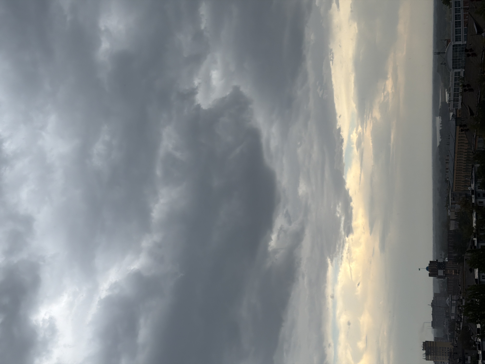 The image shows an overcast sky above a city. The clouds are dark and heavy, with some lighter patches indicating breaks in the cloud cover. Below the sky is a city skyline with various buildings, including a tower with a flag on top, and some industrial structures with smoke plumes. In the distance, a body of water can be seen, likely the Irtysh River, which flows through Pavlodar, Kazakhstan. The overall impression is a somewhat somber or dramatic atmosphere due to the heavy clouds.