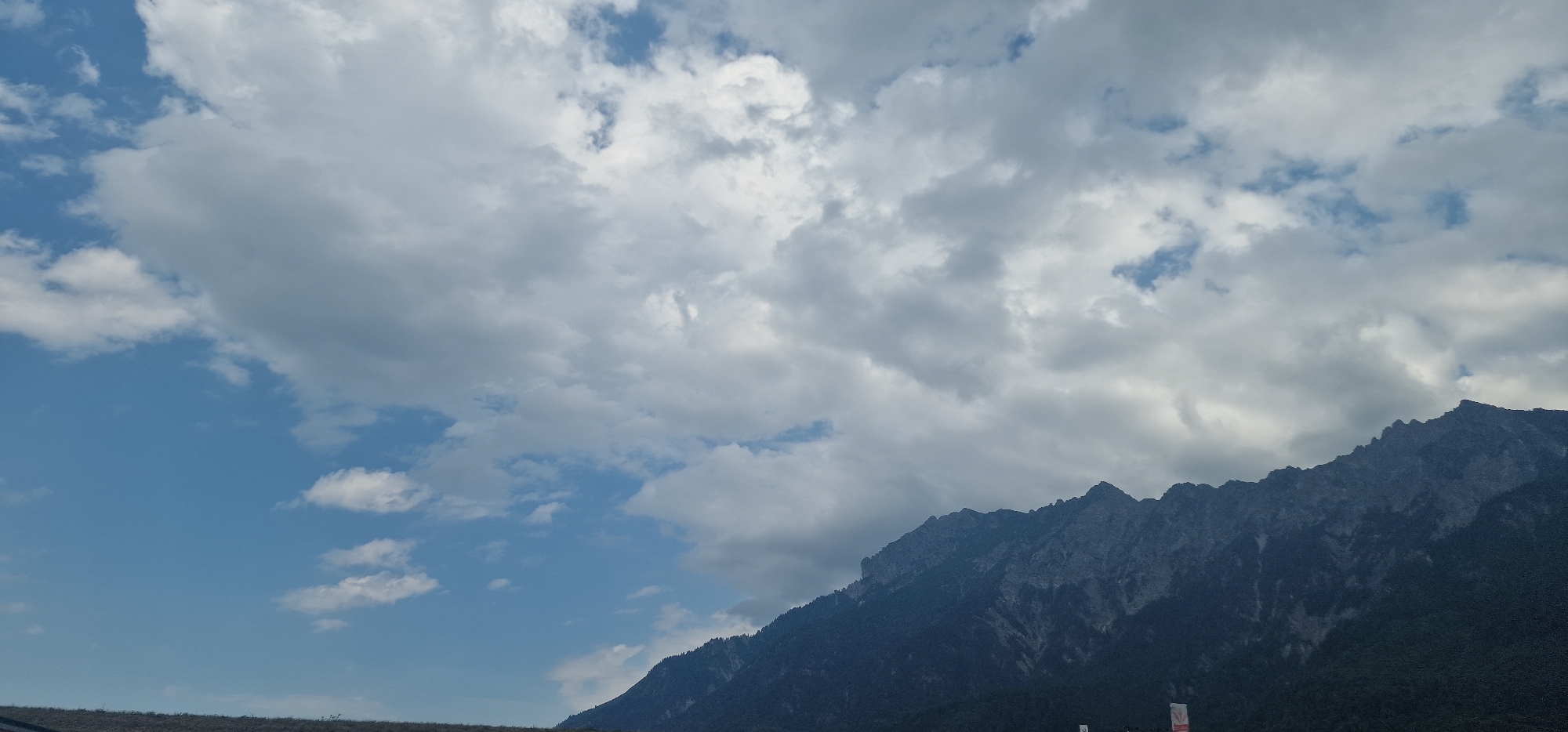 The image shows a dramatic mountain landscape under a cloudy sky. The mountains dominate the lower right portion of the frame, their jagged peaks silhouetted against the sky. Dark green foliage covers the lower slopes. The sky is a mix of bright blue patches and heavy gray clouds, suggesting an overcast or changing weather condition. In the foreground, a dry, grassy slope runs along the bottom of the image, leading up to the mountains. There is no indication of people or specific city landmarks in this view, making it difficult to ascertain the exact location within Rans, Switzerland, beyond a general mountainous terrain.