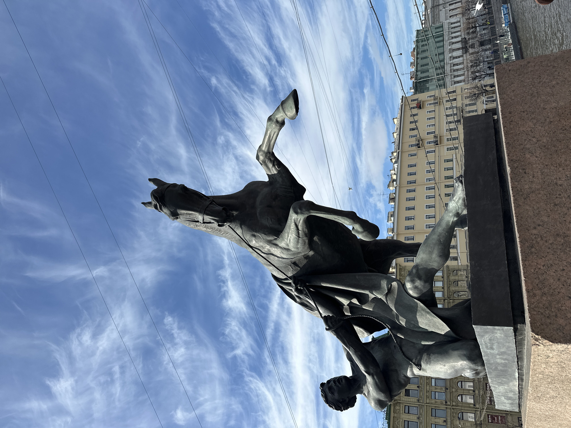 The image captures a dramatic bronze sculpture set against a bright blue sky with wispy white clouds. The central subject is a powerful horse rearing up on its hind legs, with its front legs lifted high into the air. Beneath the horse, a muscular male figure struggles with the animal, holding tightly to its reins with both hands. The man's body is tensed, conveying intense effort to control the powerful beast. Both the horse and the man are rendered in dark bronze, showcasing detailed musculature and dynamic poses.
The statue rests on a dark, rectangular base, which is placed on a reddish-brown granite pedestal visible on the right side of the frame. In the background, on the right, several multi-story buildings with classical architectural elements are visible, typical of Saint Petersburg. One building is partially covered with green construction netting or scaffolding. Beyond the buildings, a body of water, likely one of the city's canals or rivers, can be seen reflecting the sky. Thin, dark overhead lines, possibly power lines or tram wires, crisscross the sky above the statue. The scene is brightly lit by daylight, indicating a clear, sunny day. No discernible text is visible on the statue or in the background.