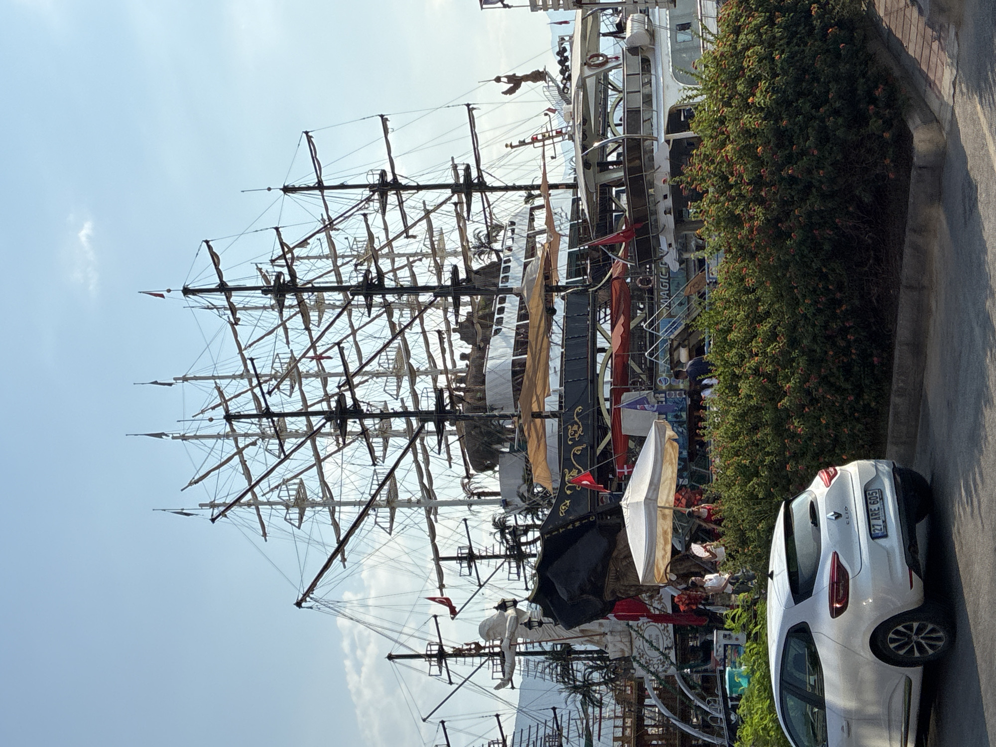 The image shows a large, multi-masted sailing ship docked in Alanya, Turkey. The ship has a complex rigging system and several decks. In the foreground, a white Renault Clio car is parked on the left. A dense green hedge with red and orange flowers runs along the edge of the parking area. Behind the hedge, a portion of the ship is visible, including a statue of a person on the bow. People are seen on the ship, some standing near a white umbrella. The sky is blue with some scattered clouds. The overall scene suggests a tourist area in Alanya, likely a harbor or waterfront promenade.