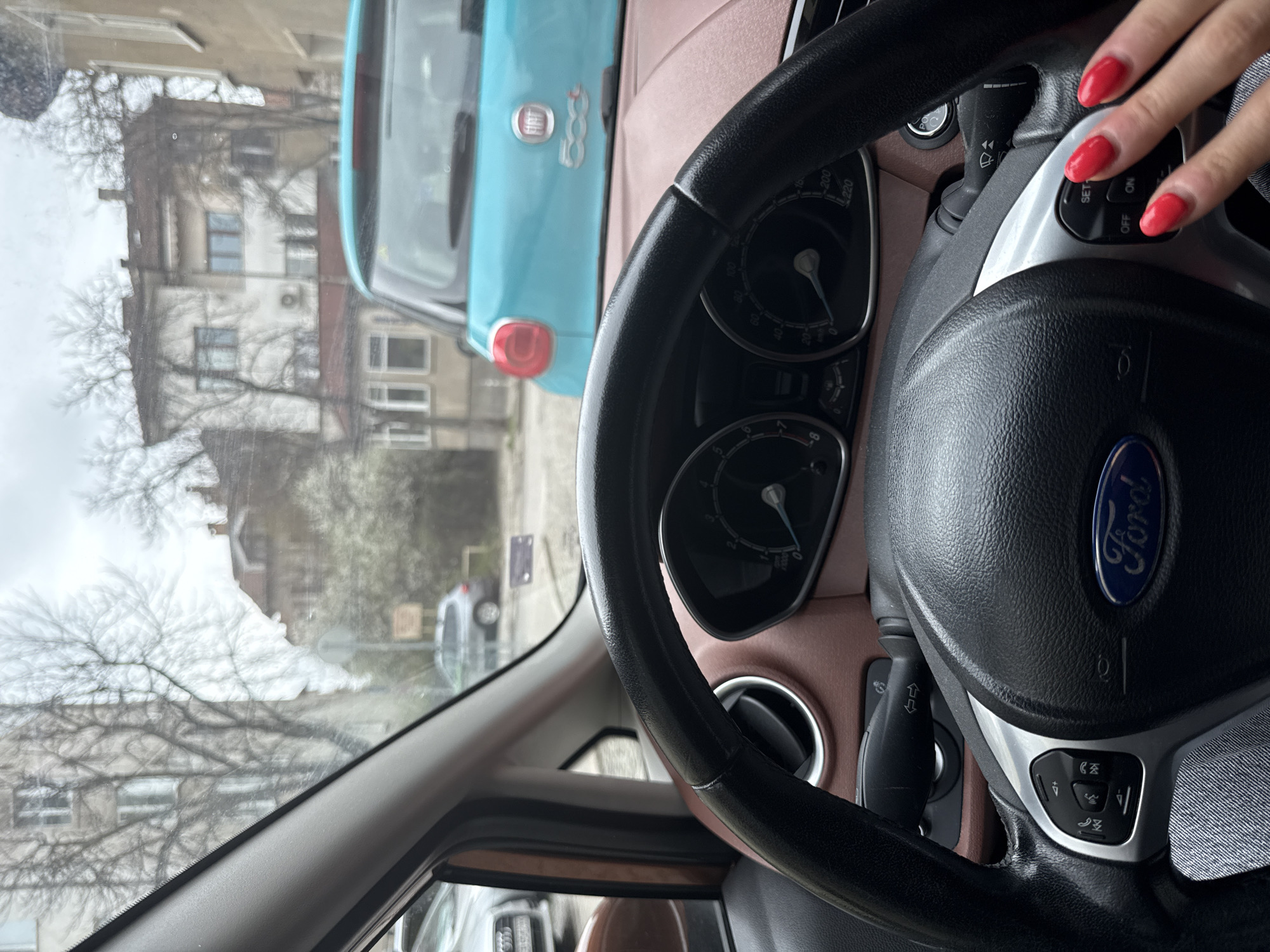 The scene is captured from inside a Ford vehicle, looking out onto a street in Sofia, Bulgaria. The dashboard and steering wheel, complete with the Ford logo, dominate the foreground. A hand with red nail polish rests on the steering wheel's control panel. Outside, a light blue Fiat 500 is visible, parked ahead. Buildings and trees line the street, suggesting an urban residential area. The image conveys a sense of daily life in a European city, observed from the driver's perspective.