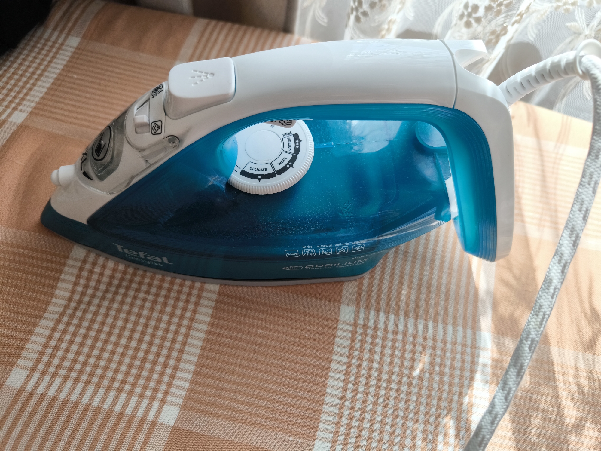 An iron is on an orange and white checkered surface. The iron is white and blue and has a dial on top with settings like "DELICATE," "WOOL," and "COTTON." The power cord is white and coils away from the iron. There is a sheer curtain in the background. The scene is indoors, possibly in Gal’chino, Russia.