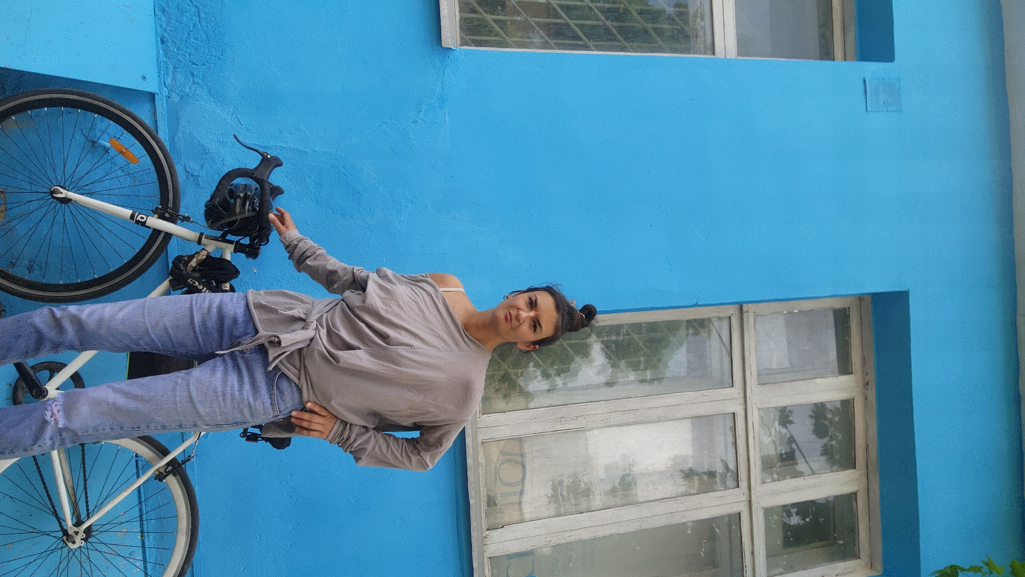 A woman in a grey long-sleeved shirt and jeans poses with a white bicycle leaning against a bright blue wall. The bicycle's front wheel is visible on the left, along with parts of its frame. Behind the woman, there are windows with white frames set into the blue wall. The location is possibly in Moscow, Russia, though there are no specific landmarks or signs visible in the image to confirm this. The woman's hair is pulled back in a bun.