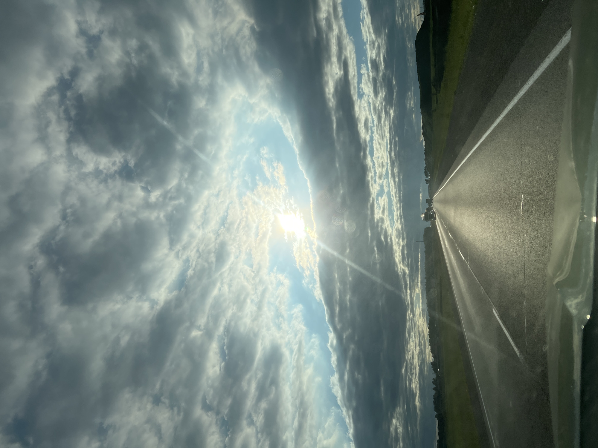 The image depicts a view from inside a vehicle looking forward onto a two-lane asphalt road. The road stretches towards the horizon, flanked by green fields on both sides. The sky is filled with dramatic, heavy clouds, with the sun breaking through in the upper center, casting bright rays of light. A few vehicles are visible in the distance on the road. The overall scene suggests a drive through a rural or semi-rural landscape, possibly during late afternoon or early evening, given the lighting. There is no specific indication of the city of Сухая Балка, Russia, in this image.