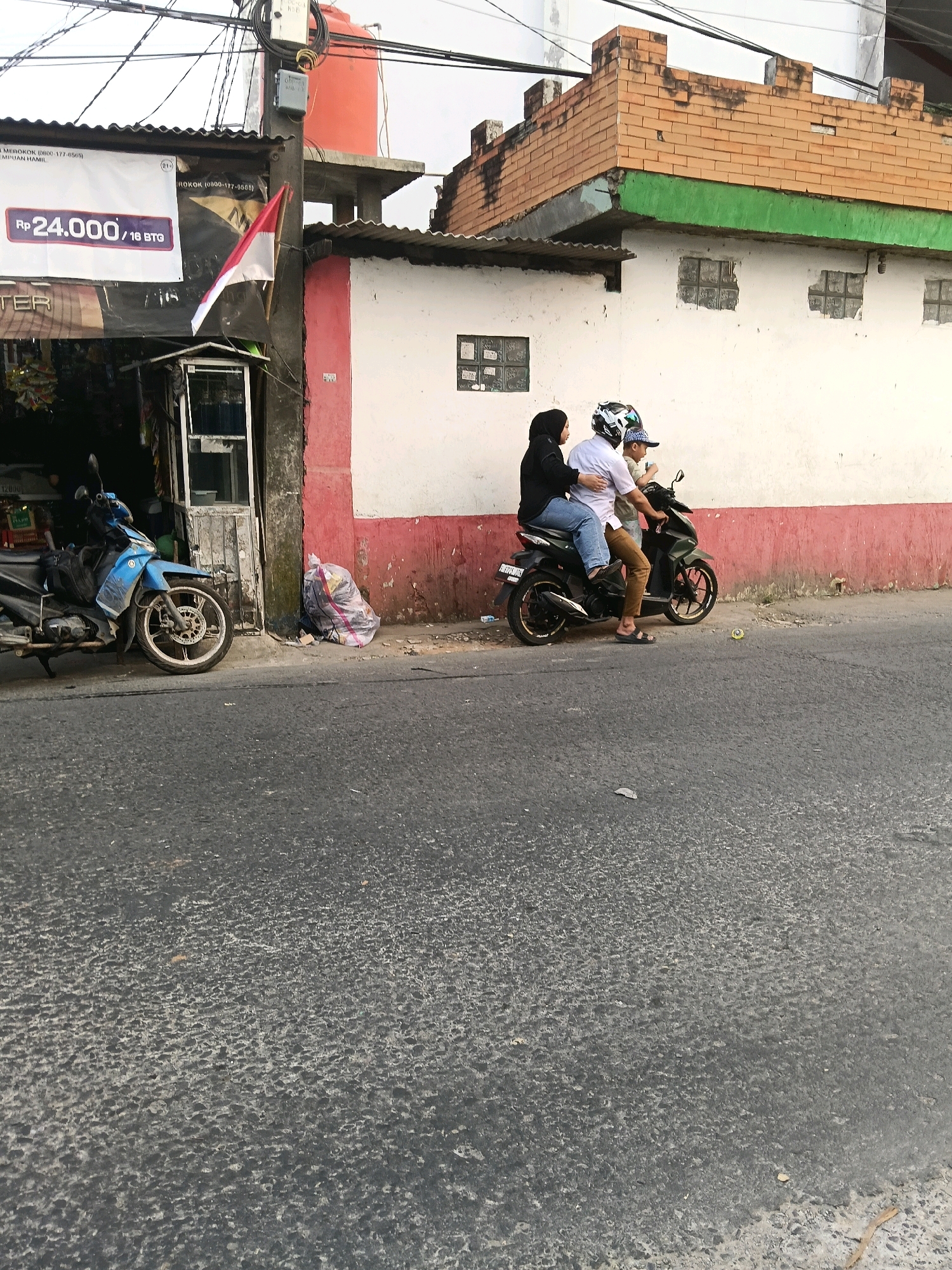 In the city of Belimbing, Indonesia, on a cloudy day, a woman wearing a black hijab and a man wearing a white shirt and blue jeans ride on a dark green motorcycle. The woman is seated behind the man and is holding onto his waist. The man is wearing a helmet with a white and black design. They are traveling on a paved road in front of a white wall with a red stripe near the bottom. To the left of the wall, a blue motorcycle is parked in front of a storefront. The storefront has a corrugated metal roof and a sign that displays "Rp 24.000/18 BTQ". A white plastic bag filled with items is leaning against the wall to the right of the storefront. In the background, a brick building with a green trim and decorative brickwork along its roofline can be seen. Electrical wires are strung across the top of the image.