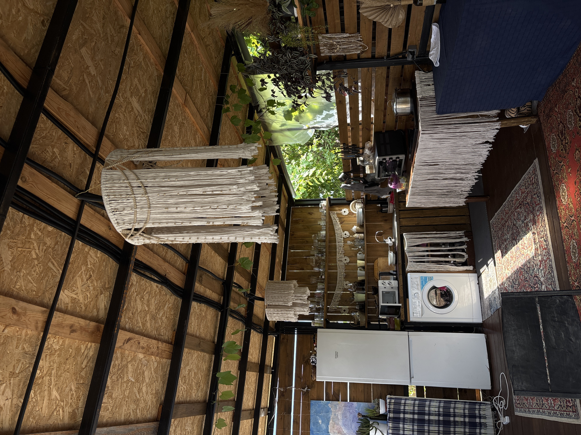The image shows an outdoor kitchen area with a rustic, bohemian design. The ceiling is made of exposed wooden beams and OSB panels, with black metal supports and electrical conduits visible. Macrame chandeliers hang from the ceiling. The walls are constructed from wooden planks.
On the left, a white refrigerator stands next to a washing machine. Above the washing machine, a microwave is placed on a wooden countertop. To the right of the microwave is a sink with a faucet. A wooden shelf unit above the countertop is filled with glassware and decorative items.
In the center of the image, a wooden table is covered with a white macrame fringe, with a black oven or toaster oven on top. To the right of this table, a dark blue rectangular object, possibly a table or cabinet, is covered with a dark blue tablecloth.
The floor is covered with several Persian-style rugs. Sunlight streams into the space, casting shadows. Lush green foliage is visible through an open section at the back of the structure, suggesting it is located in an outdoor or semi-outdoor setting surrounded by nature.
The specific location in Gudauta, Abkhazia, is not discernible from the visual information provided in the image. No people are present in the scene.