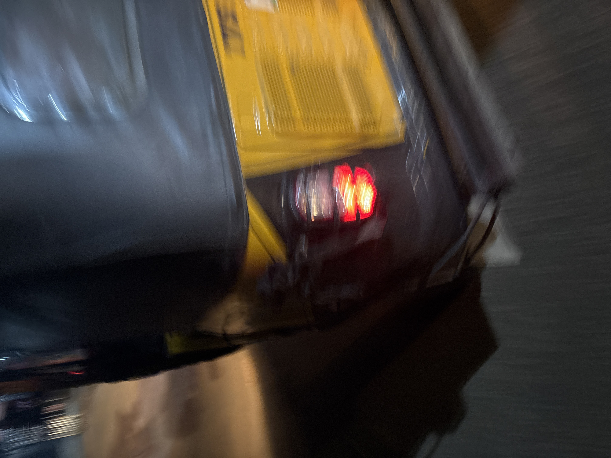 This is a blurred, low-angle, vertical shot of the rear of a yellow auto-rickshaw parked on a street at night in Onitsha, Nigeria. The auto-rickshaw has a dark canopy and a yellow panel with what appears to be a sticker or label. A red taillight is visible on the left side of the rear, illuminated and blurred due to motion or low light. The ground beneath the vehicle appears to be asphalt, with streaks of light suggesting streetlights or vehicle headlights. The overall impression is one of movement and low light conditions.