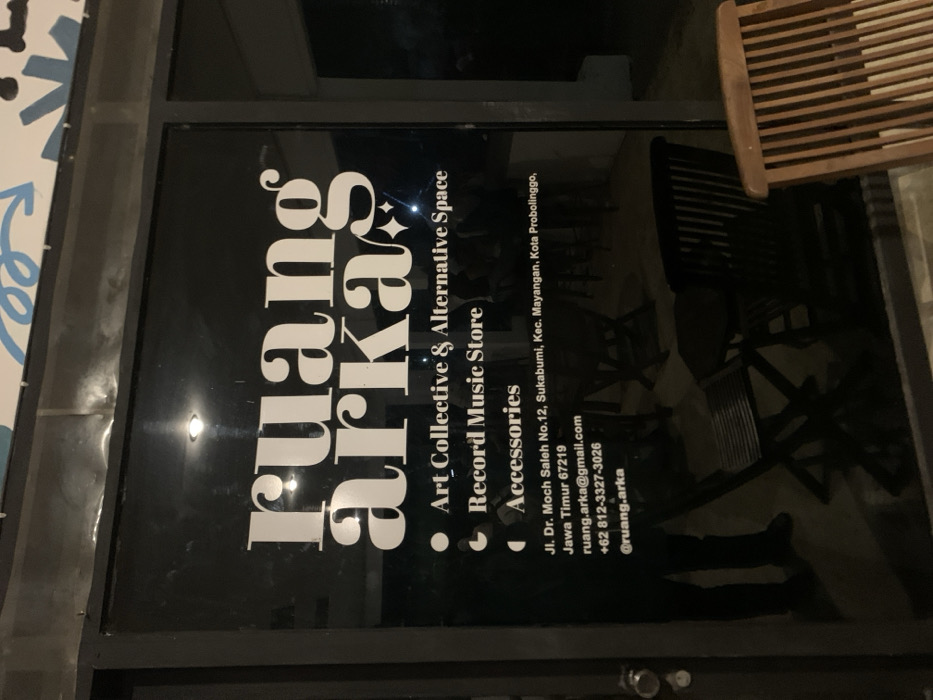 The image shows the front entrance to "ruang arka", an "Art Collective & Alternative Space" located at Jl. Dr. Moch Saleh No.12, Sukabumi, Kec. Mayangan, Kota Probolinggo, Jawa Timur 67219. The storefront window is glass and features white lettering and a black background. There is a black door to the right of the window. The window is reflective and shows the interior of the space. The interior is dimly lit and contains a few people, likely patrons of the space. There are two wooden chairs in the foreground. The space also appears to be a "Record Music Store" and sells "accessories". There is a social media handle displayed on the window: "@ruang.arka".