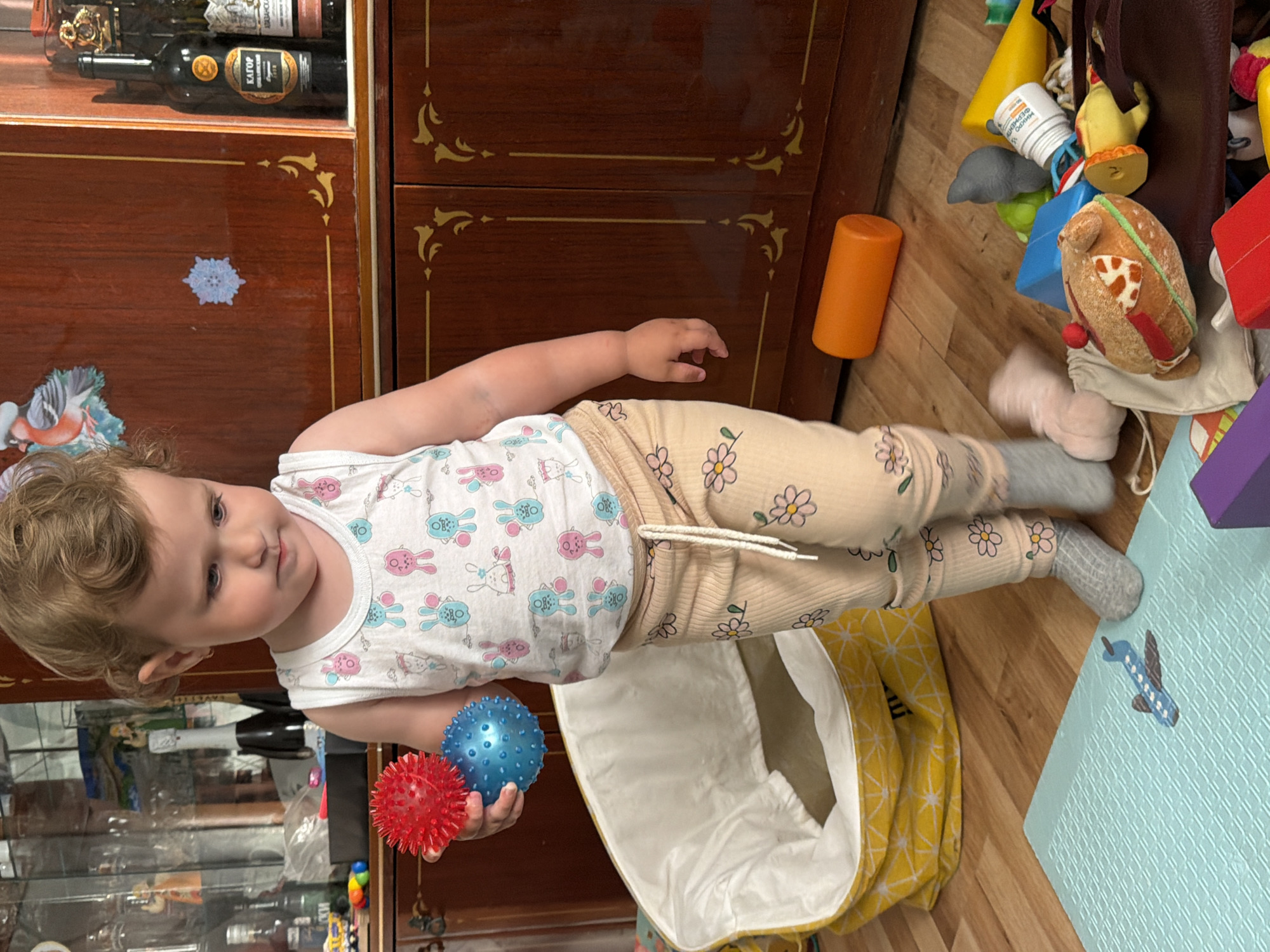 A toddler stands on a wooden floor in front of a wooden cabinet. The toddler is wearing a white tank top with cartoon rabbits and beige pants with a floral pattern, and grey socks. They are holding a red and blue textured ball in their left hand. To the left of the toddler is a large, yellow and white patterned laundry basket. Scattered on the floor are various toys, including a blue airplane toy on a light blue mat, an orange cylinder, a stuffed hamburger, and other colorful objects. The wooden cabinet behind the toddler has decorative gold trim and appears to be part of a room. The scene is indoors, in what looks like a living area or playroom. The location is Komsomolskiy, Russia.