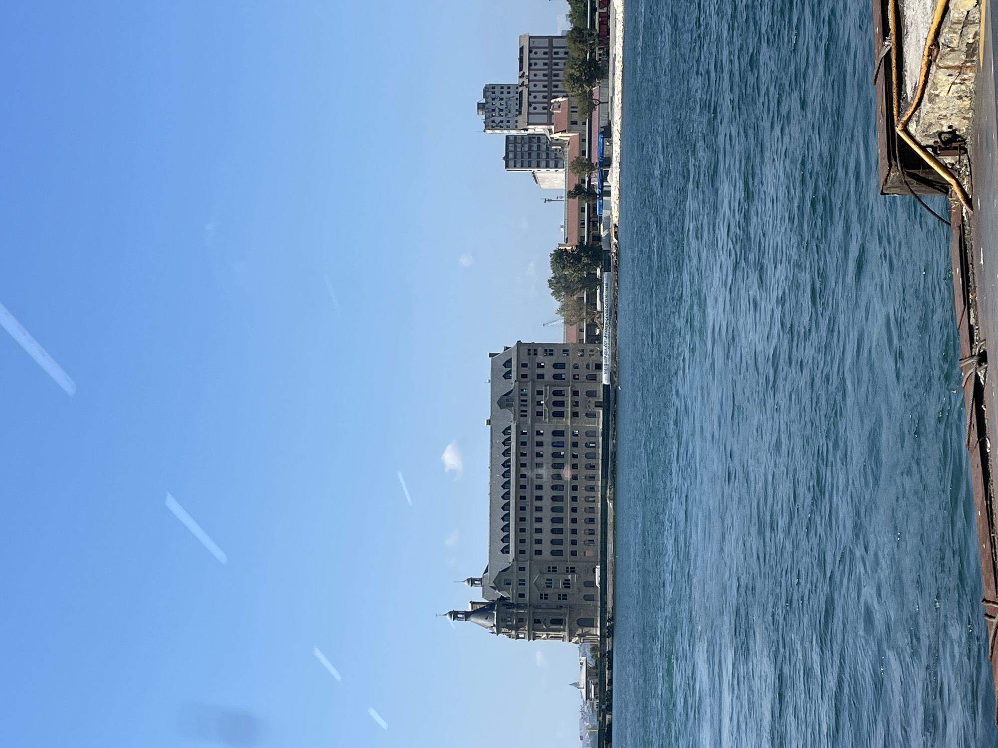 The image shows a view from a body of water, likely the Bosphorus Strait, looking towards the Kadıköy district of Istanbul, Turkey. In the foreground, a rusty metal structure is partially visible, suggesting the presence of a dock or a ferry. The water is a deep blue with visible ripples. Dominating the middle ground is the Haydarpaşa Terminal, a large, historic railway station with distinctive architecture, including a prominent clock tower. To the right of the terminal, other buildings of varying heights and styles are visible along the coastline, some appearing to be industrial or commercial. Trees and vegetation are interspersed between the buildings. The sky is clear and blue with a few faint white streaks, possibly contrails from aircraft. No people are visible in the frame.