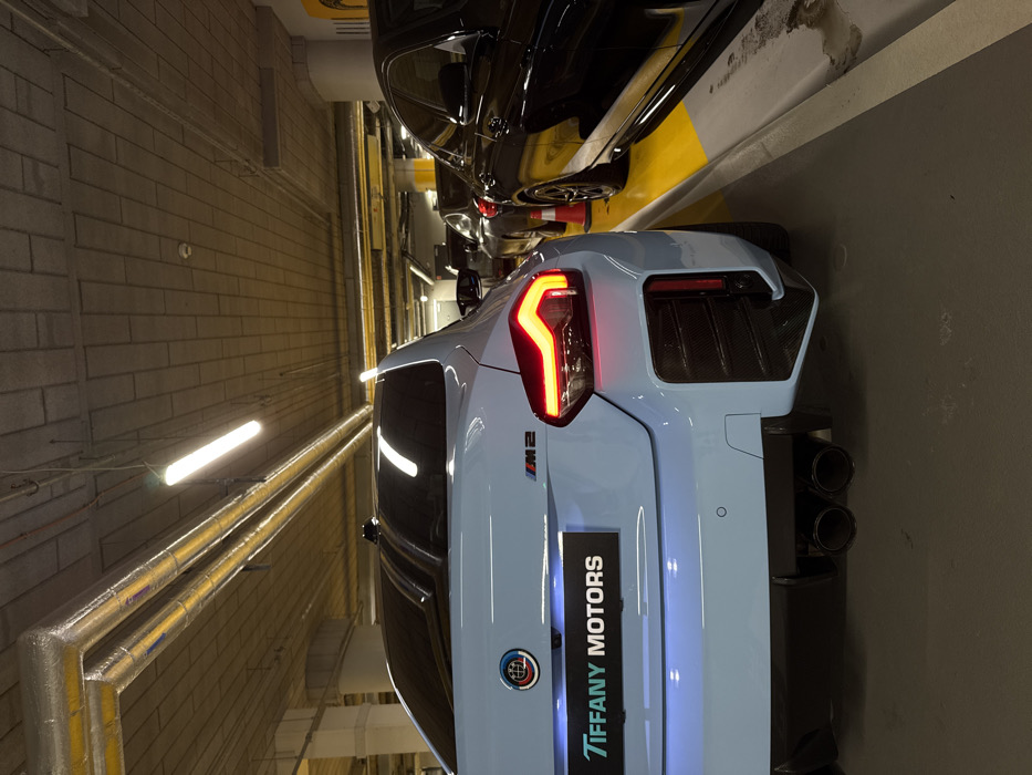 A light blue 2023+ BMW M2 (G87) coupe is prominently parked in an underground parking garage, viewed from its rear-left quarter. The vehicle's left taillight is illuminated red. The trunk lid features a modified BMW emblem with a blue and red globe design, and "M2" badging. A black panel below the trunk displays "TIFFANY MOTORS" in light blue text. The rear bumper incorporates carbon fiber-look trim on the side vents and a dark diffuser housing twin black exhaust tips.
To the immediate right of the BMW M2, a black sedan is parked behind a yellow-painted curb or barrier, with a small orange traffic cone positioned between the two vehicles. Additional parked cars are visible in the background. The environment consists of a concrete ceiling with visible rectangular panels, exposed metallic ventilation ducts, some wrapped in yellow insulation, and overhead fluorescent tube lighting. The floor is dark grey concrete with yellow parking space markings. No individuals are present. This scene is located at an underground parking facility, specifically identifiable as being associated with Tiffany Motors at ul. Giełdowa 1, 01-211 Warsaw, Poland.