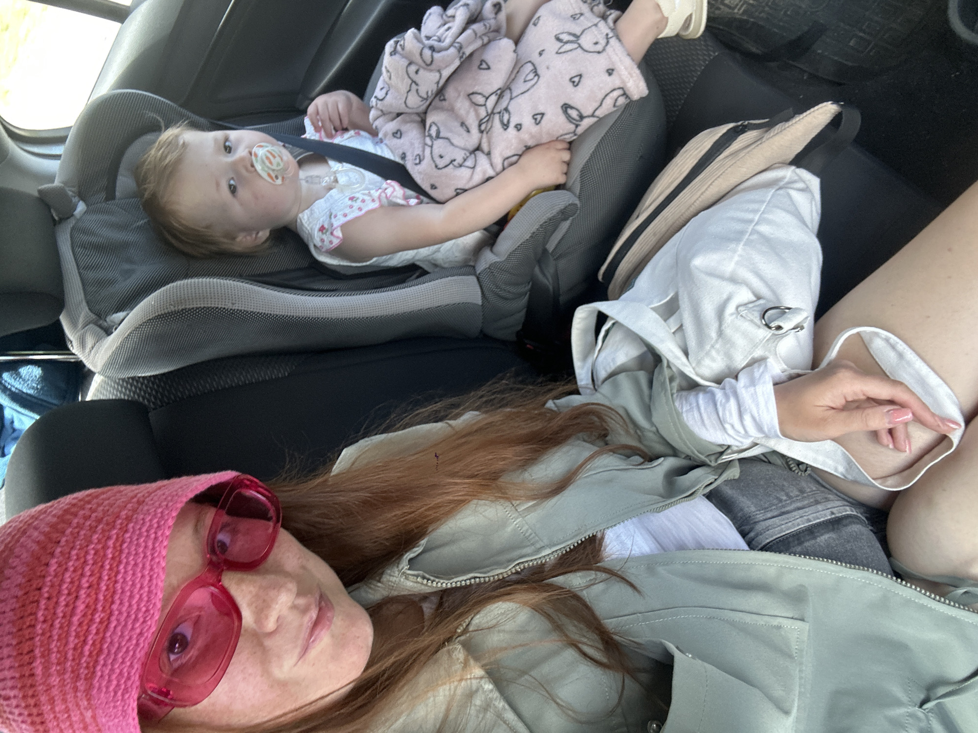 This image, taken in Nizhny Novgorod, Russia, presents a top-down view inside a car, featuring an adult and a young child.
In the foreground, an adult, presumably female, is seen reclining or lying down across a car seat. She has reddish-brown hair and is wearing a bright pink knitted beanie and large, rectangular pink-framed sunglasses, which obscure most of her eye area but reveal a relaxed expression and some freckles on her face. She is dressed in a light green zip-up jacket over a white long-sleeved top and dark grey or black shorts/skirt. Her hand, featuring light pink manicured fingernails, is visible near her leg.
Further back, a young child, appearing to be a toddler, is secured in a grey car seat. The child has light brown hair and is wearing a light-colored top with a subtle pattern. A white pacifier with a colorful design is in their mouth, and they are looking directly towards the camera. A pink blanket, adorned with white hearts and rabbit patterns, covers the child's legs and lower body.
The car's interior features dark upholstery and floor mats. Next to the adult's legs and below the child's car seat, there is a white canvas-like bag with a metal D-ring, and a beige corduroy fanny pack or small bag. The lighting indicates it is daytime. The scene suggests a moment of rest or travel. No visible text is present in the image.