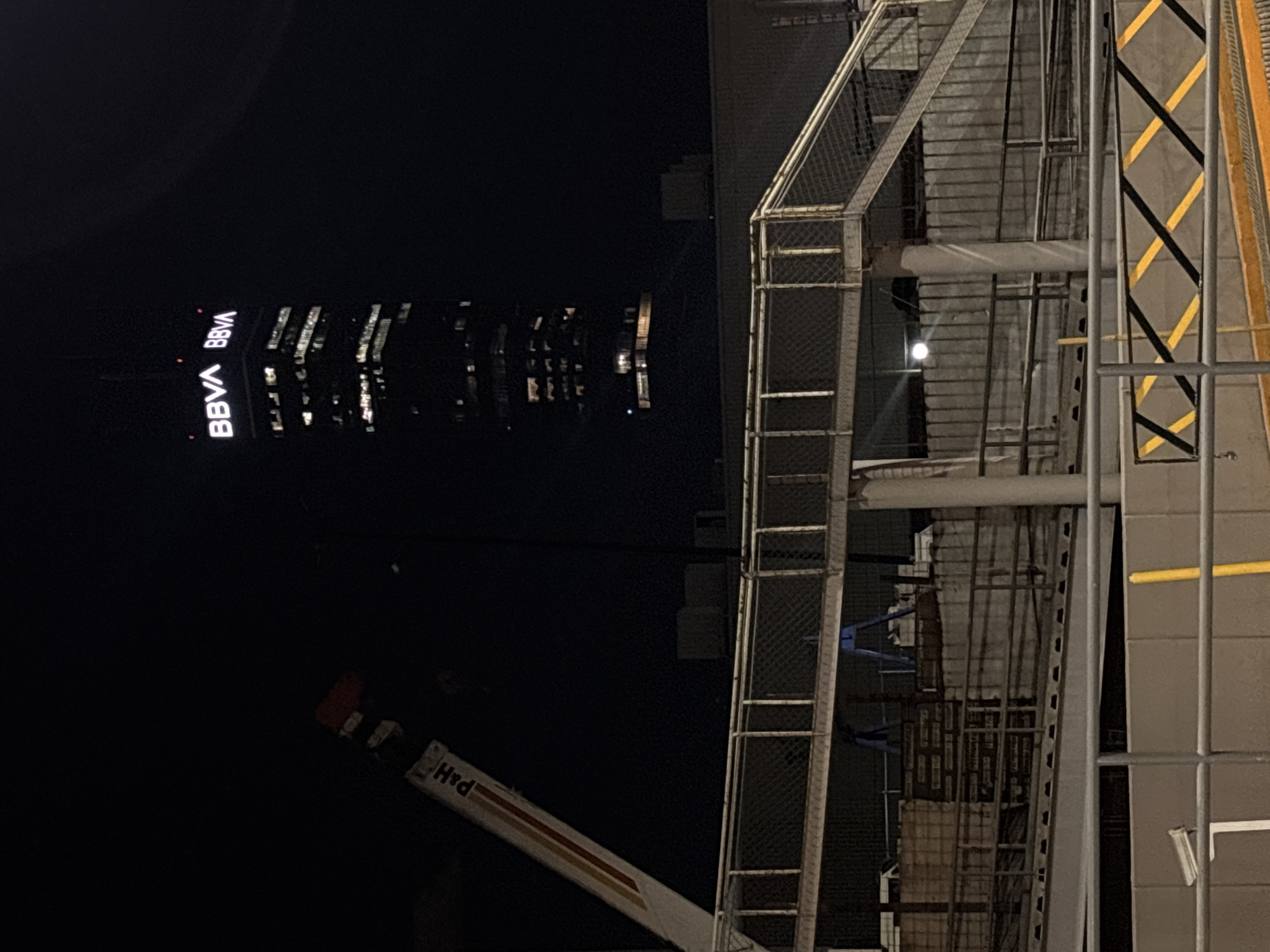 The image, taken at night in San Bernardino Tlaxcalancingo, Mexico, shows the BBVA building illuminated in the distance. In the foreground, there is construction scaffolding and part of a crane. The crane's boom bears the text "H+D". The scaffold is made of metal and has a cage-like structure. A wall or structure adjacent to the scaffolding has diagonal stripes of yellow and black. The lighting conditions suggest a nighttime environment with artificial illumination from the building and potentially nearby sources.
