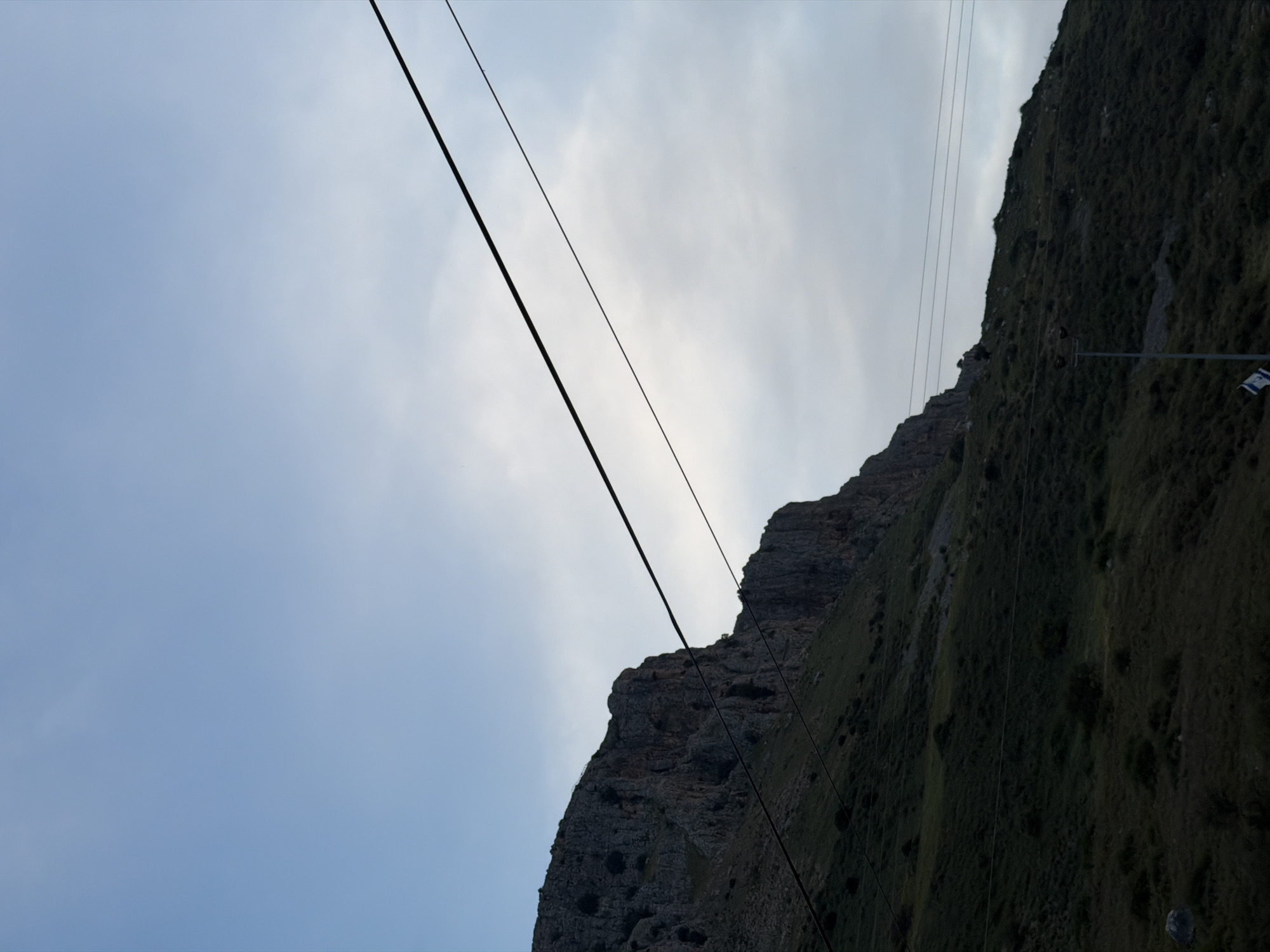 The image displays a steep, rugged mountain slope against a cloudy sky, likely captured from an ascending cable car. The sky, occupying the upper-left portion, exhibits pale blue tones blending into brighter white cloud cover towards the right, suggesting an overcast or partly cloudy day with the sun potentially obscured.
The lower-right of the image is dominated by the mountain, which features distinct rocky outcrops and is heavily covered in dark green vegetation, including grass and shrubs. Multiple dark cables crisscross the scene; two thicker, prominent cables run diagonally across the sky and mountain, indicative of a cable car system. Several thinner, parallel cables are also visible along the mountain's vegetated slope, possibly part of the lift infrastructure or power lines.
On the right side of the mountain, a vertical pole-like structure with a horizontal arm is visible. From this arm hangs a blue and white flag, clearly identifiable as the flag of Israel due to its two blue stripes and the central Star of David design, a strong location cue for Migdal, Israel.
No people are visible. The overall scene depicts a natural, mountainous landscape during daytime, viewed from a low angle, highlighting both the natural terrain and human-made transport infrastructure.