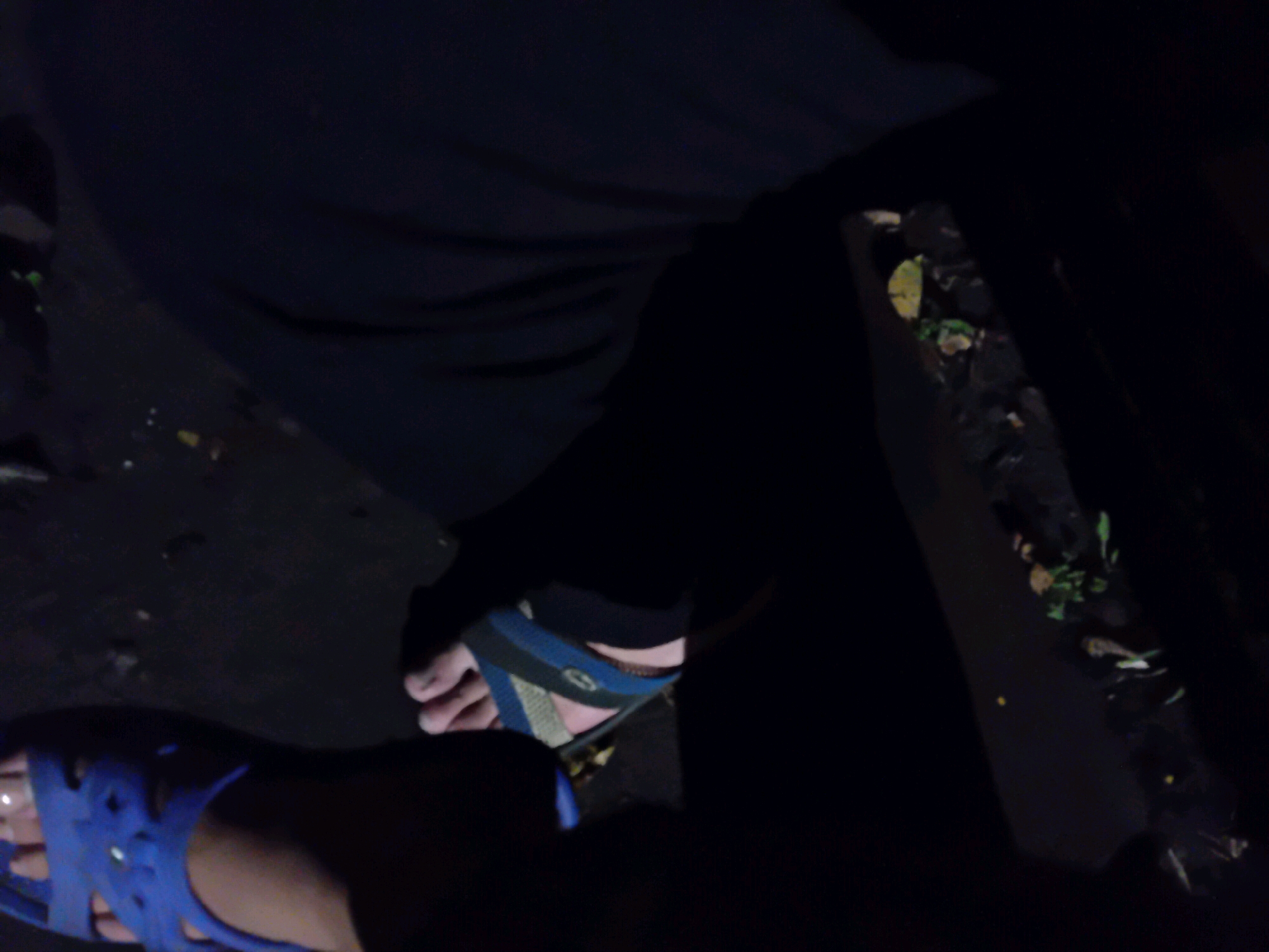 In a low-light, dark environment possibly at night, the image focuses on a person's legs and feet. The left foot is clad in a blue sandal with a strappy design. The right foot is wearing a sandal with blue and green straps. The individual is wearing dark clothing, likely pants or leggings. The ground beneath appears to be dark and possibly muddy or dirt, with scattered small leaves or debris. The overall scene suggests an outdoor setting, potentially in a less urbanized area or a poorly lit part of the city of Chuhuiv, Ukraine, given the context of the provided location. The image quality is poor due to low light conditions.