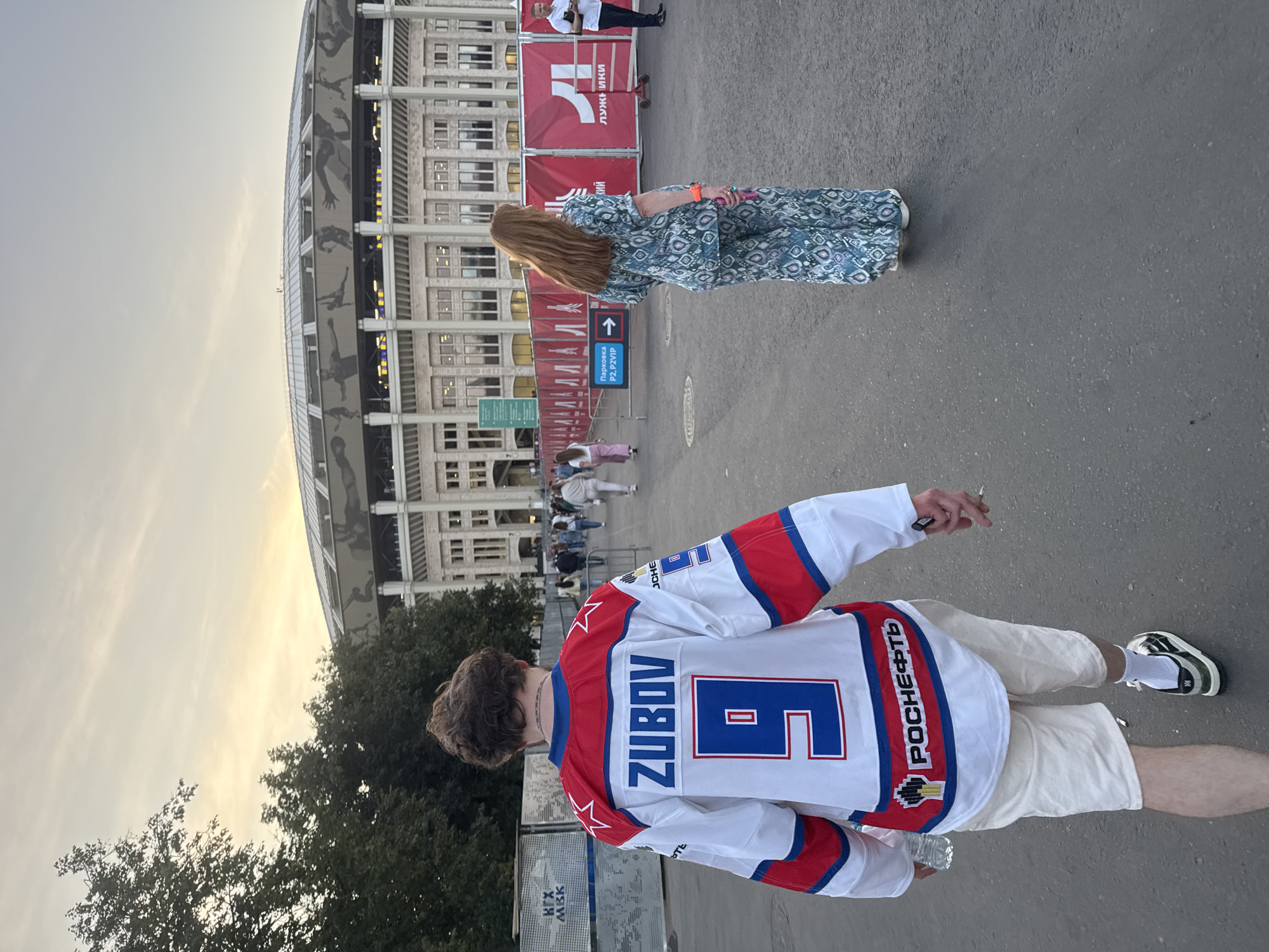 In Moscow, Russia, a person wearing a white hockey jersey with "ZUBOV" and the number "9" in blue, along with red and blue trim, walks away from the camera. They are wearing off-white shorts and sneakers. To their right, a woman in a blue and white patterned jumpsuit walks in the same direction. In the background, the Luzhniki Stadium, a large sports complex, is visible. The sky is overcast with a hint of sunset. The area appears to be an outdoor pathway leading to the stadium, with banners and fencing along the side.