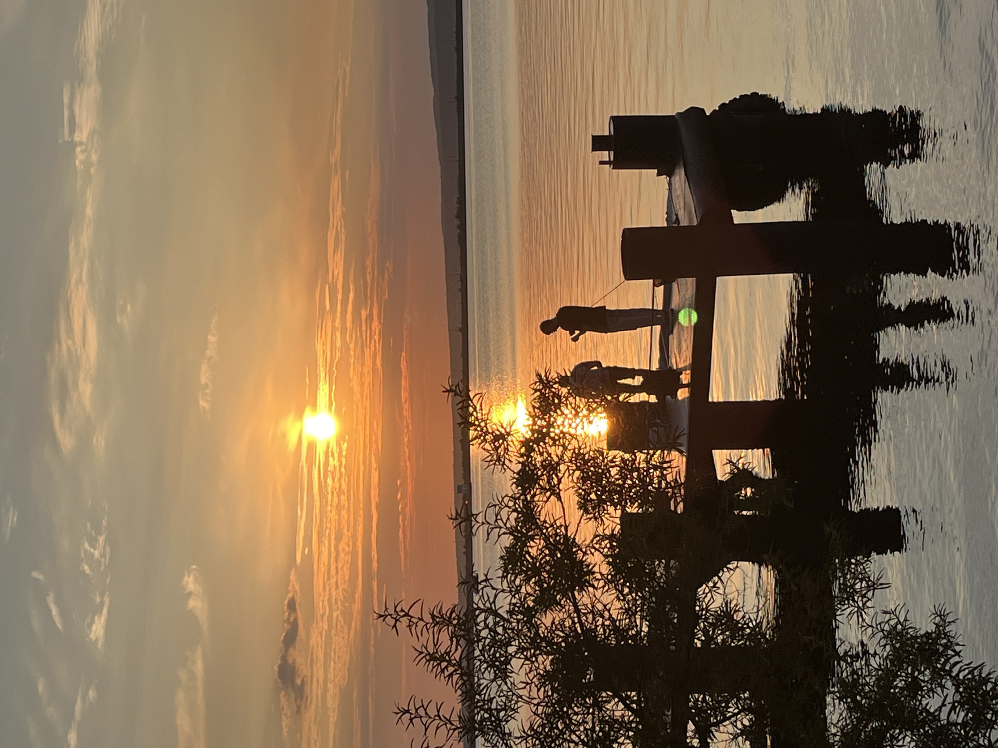 At sunset, three figures stand on a pier jutting into a calm body of water, likely the Kama River in Chaikovsky, Russia. The sun, a bright orb, is low on the horizon, casting a warm orange glow across the sky and water. Silhouetted against the vibrant sunset, two of the figures appear to be fishing, one holding a rod, while the third stands slightly apart. The pier itself is dark and partially submerged, with thick wooden posts supporting its structure. The foreground is dominated by the dark silhouette of a tree with sparse foliage, its reflection shimmering in the water alongside the pier's structure. In the distance, a hazy shoreline with industrial structures is visible.