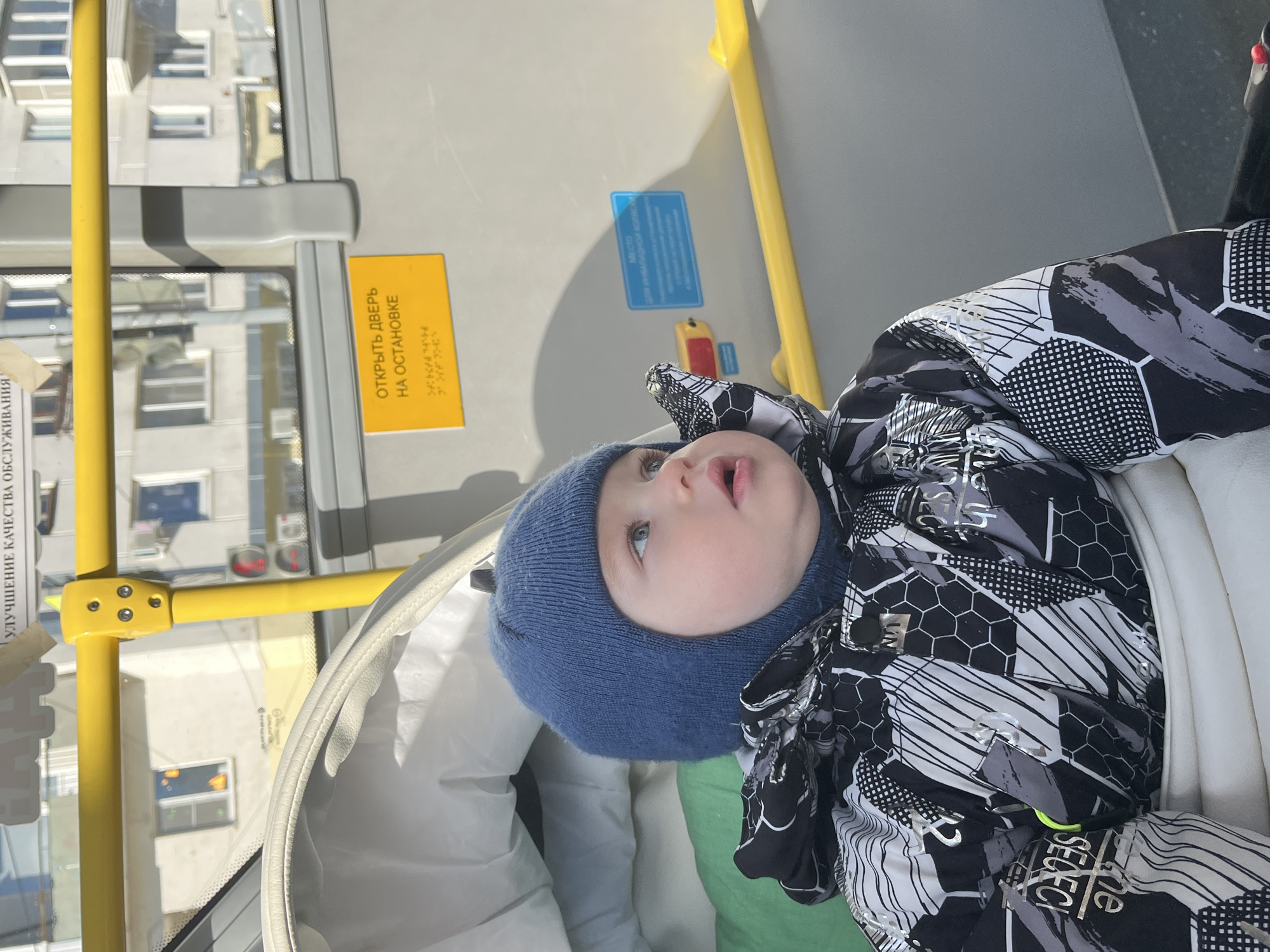 A baby wearing a blue knit hat and a black, white, and gray patterned winter jacket is resting in a stroller, presumably in a public transportation vehicle such as a bus in Chaikovsky, Russia. The baby is looking up. Yellow handrails and various signs in Russian are visible on the bus. Outside the window, several buildings are visible, indicating an urban environment. A sticker says "Improvement of Quality Service". A sign that states "Open door at stop" is also visible.