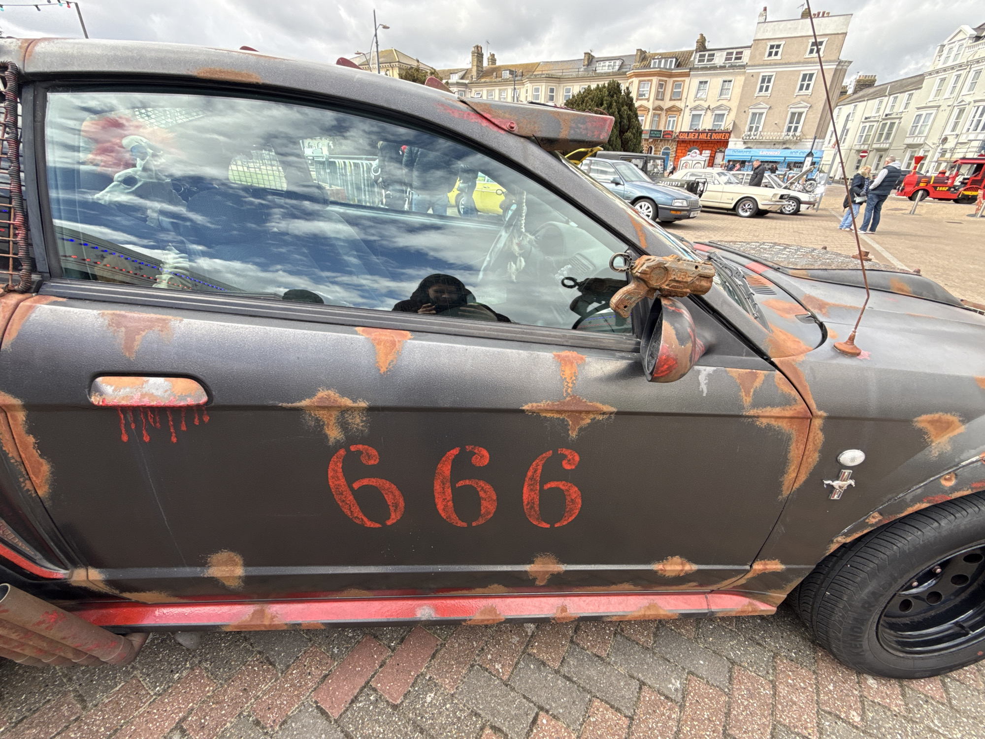The image shows a heavily modified, rust-covered car with the number "666" painted on the door. The car has a weathered appearance, with simulated rust and red paint suggesting blood splatters near the door handle. An object resembling a toy gun is attached to the side mirror. The car appears to be parked on a brick-paved area. In the background, buildings characteristic of Great Yarmouth, United Kingdom, can be seen, with a mix of architectural styles. Other vehicles and pedestrians are visible, suggesting a public event or gathering. The sky is overcast, indicating a cloudy day.