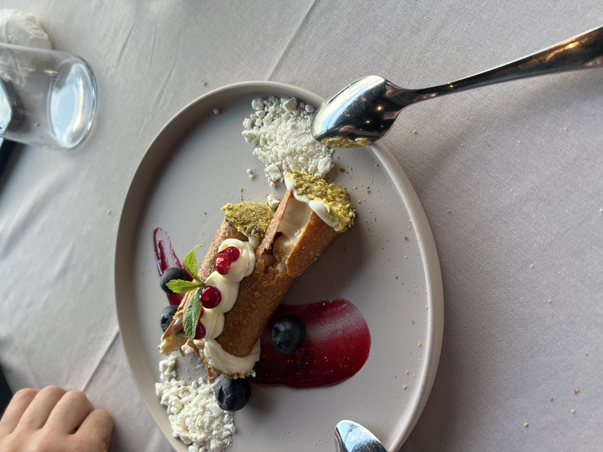 The image shows a plate of two cannoli pastries on a light gray plate. The cannoli are filled with cream and decorated with red berries, blueberries, and mint leaves. Pistachios are on the ends of the cannoli. There's a smear of red sauce under one cannoli and some crumbled white substance on the plate in two places. A spoon rests on the plate, its bowl pointing toward the cannoli. A glass of water is partially visible in the upper left of the image, and part of a hand is in the lower left of the image. The setting appears to be a restaurant table, covered with a light gray tablecloth. The location is likely in Burgas, Bulgaria, but this can't be definitively determined from the image alone.