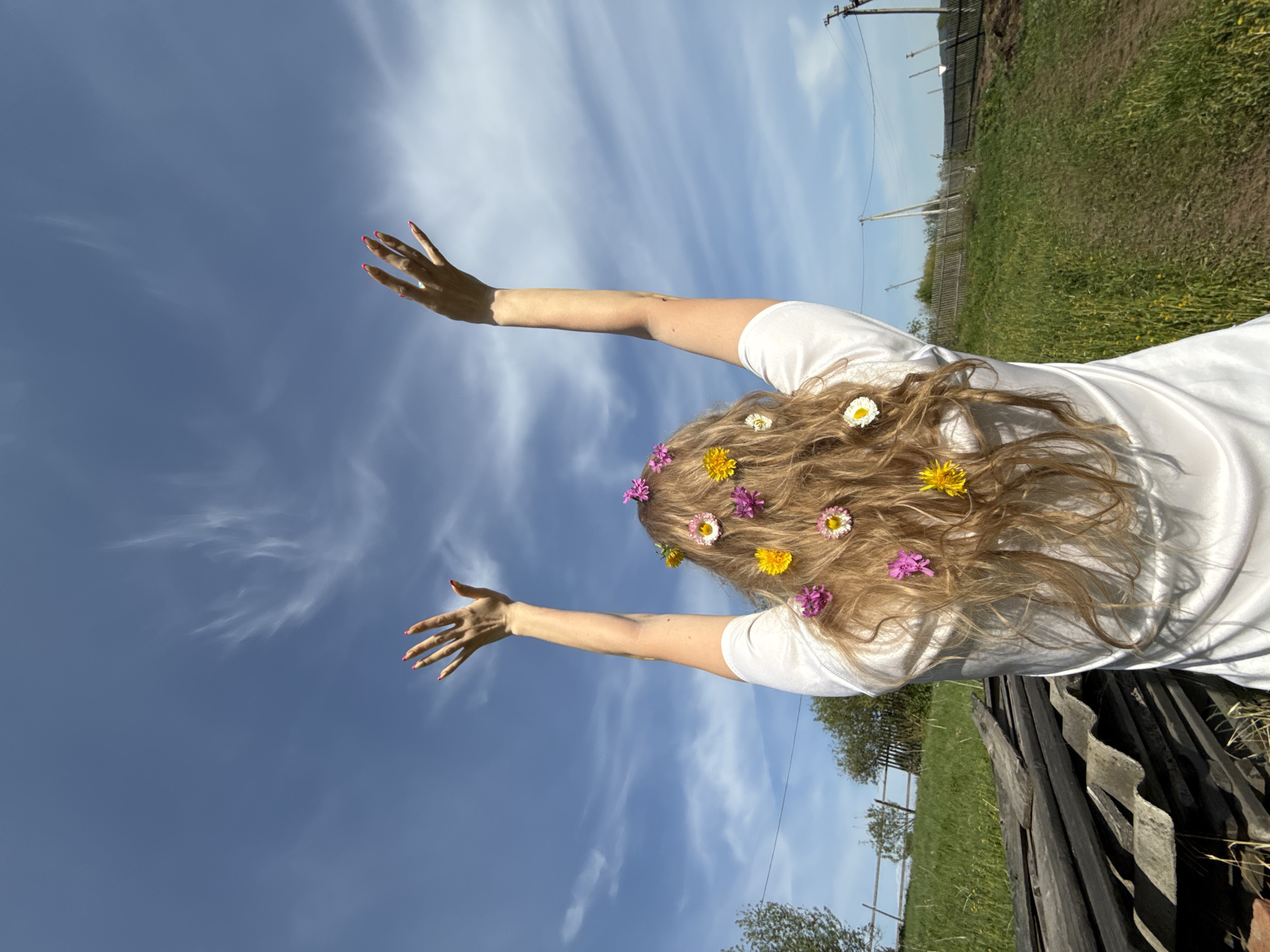A fair-skinned individual is seen from the back, lying in a grassy field under a blue sky with wispy clouds. The person wears a white t-shirt and has long, wavy blonde hair adorned with pink, yellow, and white flowers. Both arms are raised, hands open, towards the sky. The fingernails are painted pink. The field is green and slightly overgrown. In the background, a fence and some trees are visible. Also, a wooden fence is visible and what looks like a metal roof. The scene appears to be in a rural or semi-rural location, possibly near Nizhnyaya Chermoda, Russia.