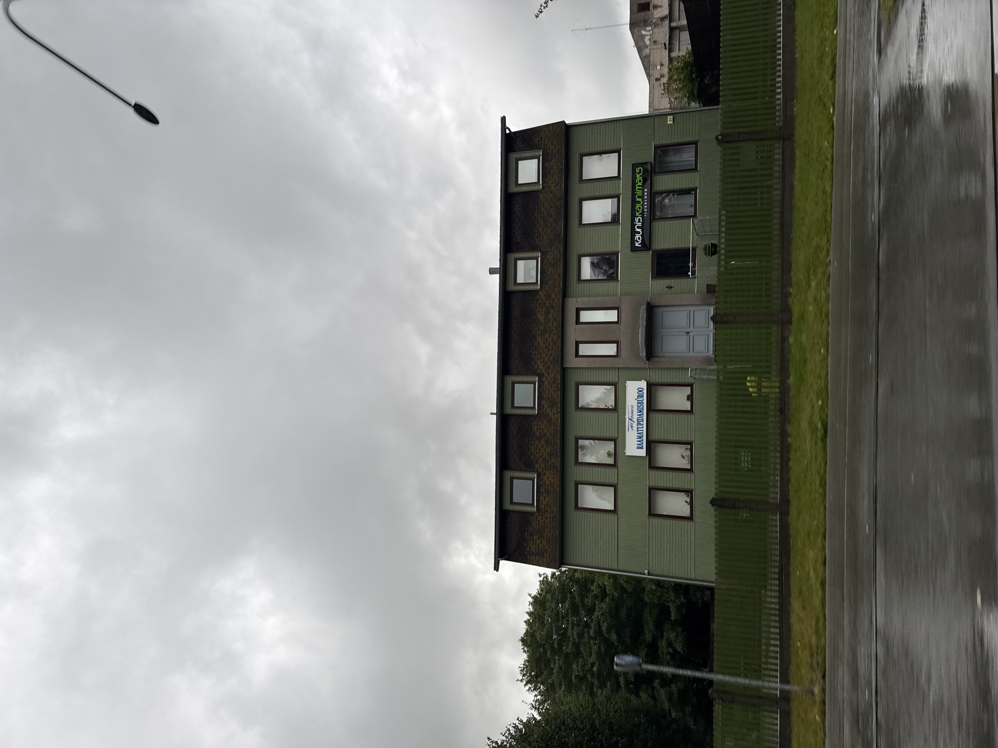 The image captures a two-story green building with a dark brown mansard roof against an overcast sky. The building has multiple windows on both stories and a prominent sign on the second story that reads "KAUNIS KAUNIMAKS ILUSALONG". Another sign on the same level says "RAAMATUPIDAMISBÜROO". There is a green wooden fence in front of the building, and a wet asphalt road in the foreground. The general scene suggests an urban or suburban environment, possibly a commercial or office building. The presence of multiple signs indicates different businesses operating within the building. The overcast sky and wet road imply recent or ongoing rainfall. The building appears to be located in Tallinn, Estonia, given the presence of Estonian language on the signs.
