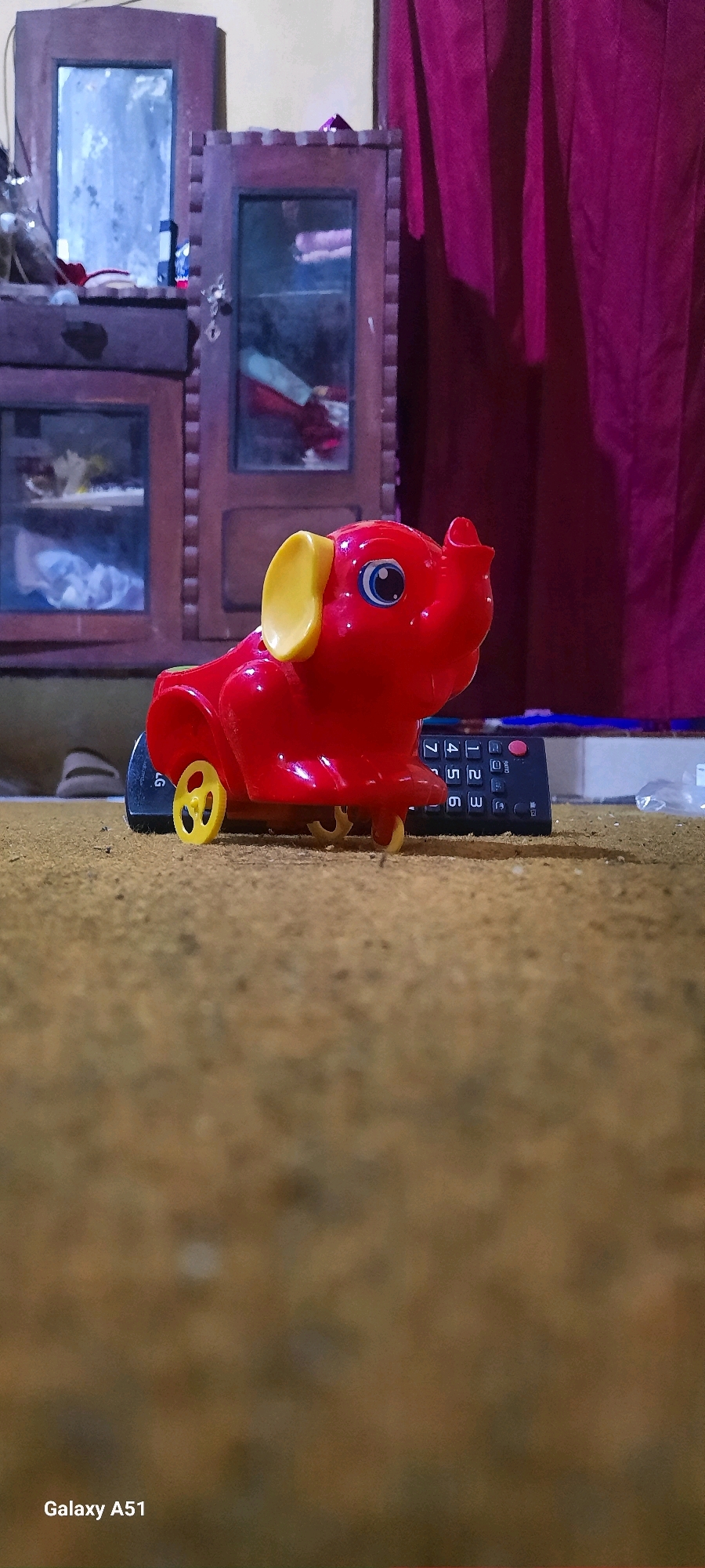 A red toy elephant with yellow ears and wheels sits on a textured surface, possibly carpet or a mat. A black remote control with numbered buttons is placed behind the toy. In the background, a dark red curtain hangs, and a wooden cabinet with glass doors is visible, reflecting light and some objects inside. The scene appears to be indoors, with a low-angle perspective. The bottom left corner of the image displays "Galaxy A51". The context of Jatitengah, Indonesia is provided.
