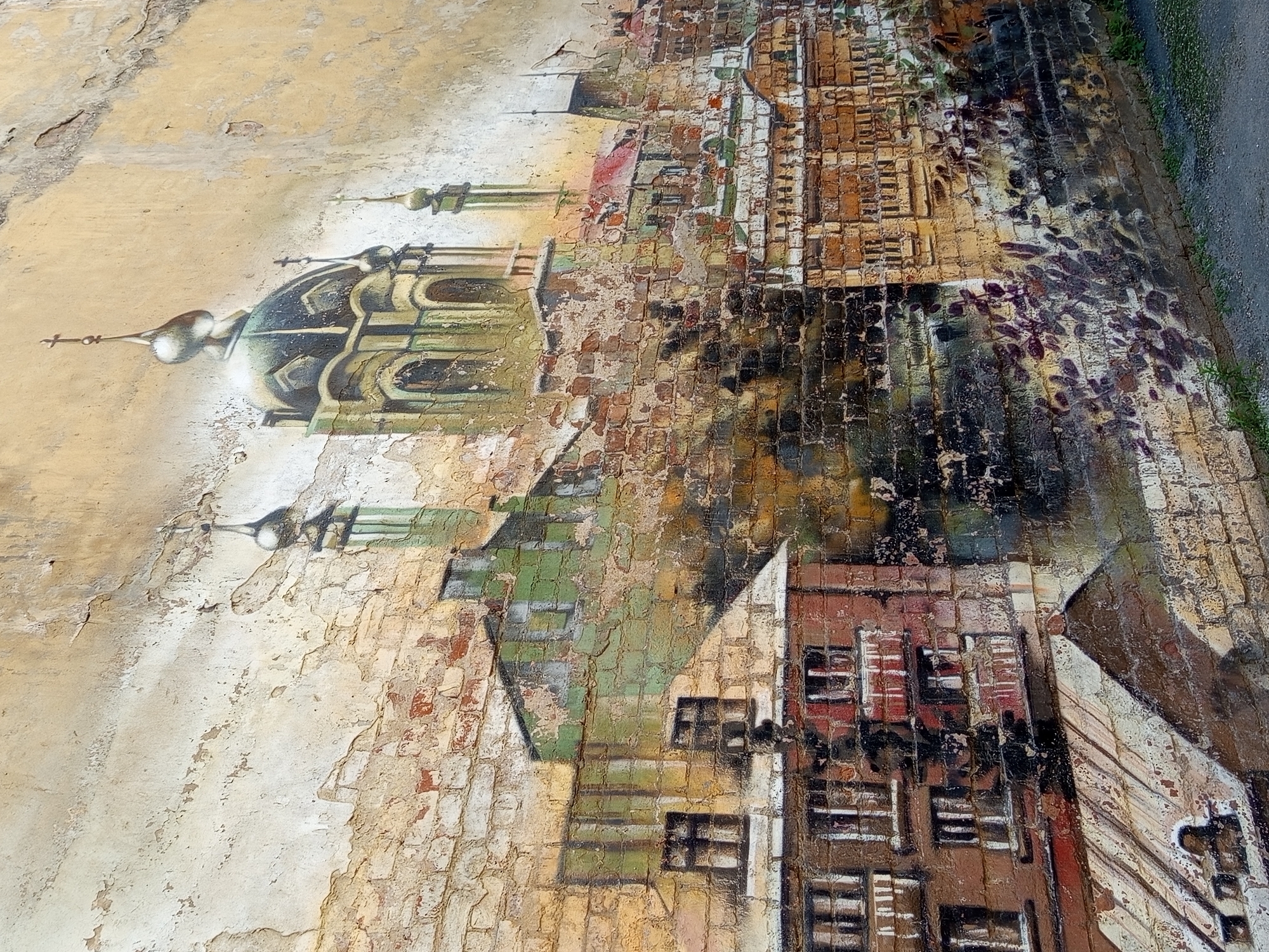 This is a photograph of a fresco on a brick wall in Saint Petersburg, Russia. The fresco depicts a cityscape with architectural elements resembling churches or cathedrals with domes and spires. In the foreground, there are buildings with windows and balconies, rendered in earthy tones and brick red. The background features the grander structures with green and gold accents. The fresco appears weathered, with parts of the paint peeling and the underlying brickwork showing through, suggesting age and exposure to the elements. The overall impression is of an old European city with historical architecture. The bottom of the image shows a glimpse of a grey asphalt surface, possibly a sidewalk or street, with some sparse green vegetation growing alongside the wall.