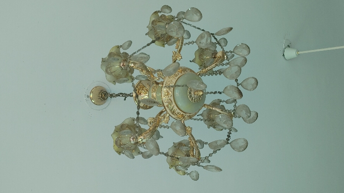 The image displays an ornate, multi-arm chandelier suspended from a light-colored ceiling. The chandelier features a central, light-toned body with intricate gold-colored decorative banding and finials. Six curved, gold-toned arms extend outwards, each supporting a light-colored, flower-shaped glass or alabaster cup. Numerous clear, faceted crystal pendants, predominantly teardrop-shaped, are suspended from the chandelier's arms and central structure by small chains.
The ceiling is a uniform light hue, possibly white or off-white, showing minor dust or discoloration around the chandelier's circular mounting plate. In the lower right portion of the frame, a separate white cylindrical ceiling fixture, possibly a light cord or fan mount, is visible, also with some discoloration at its attachment point. The scene is an interior environment, consistent with a residential or commercial building. No individuals, specific actions, or interactions are depicted. This scene is located within the city of Savar, Bangladesh.