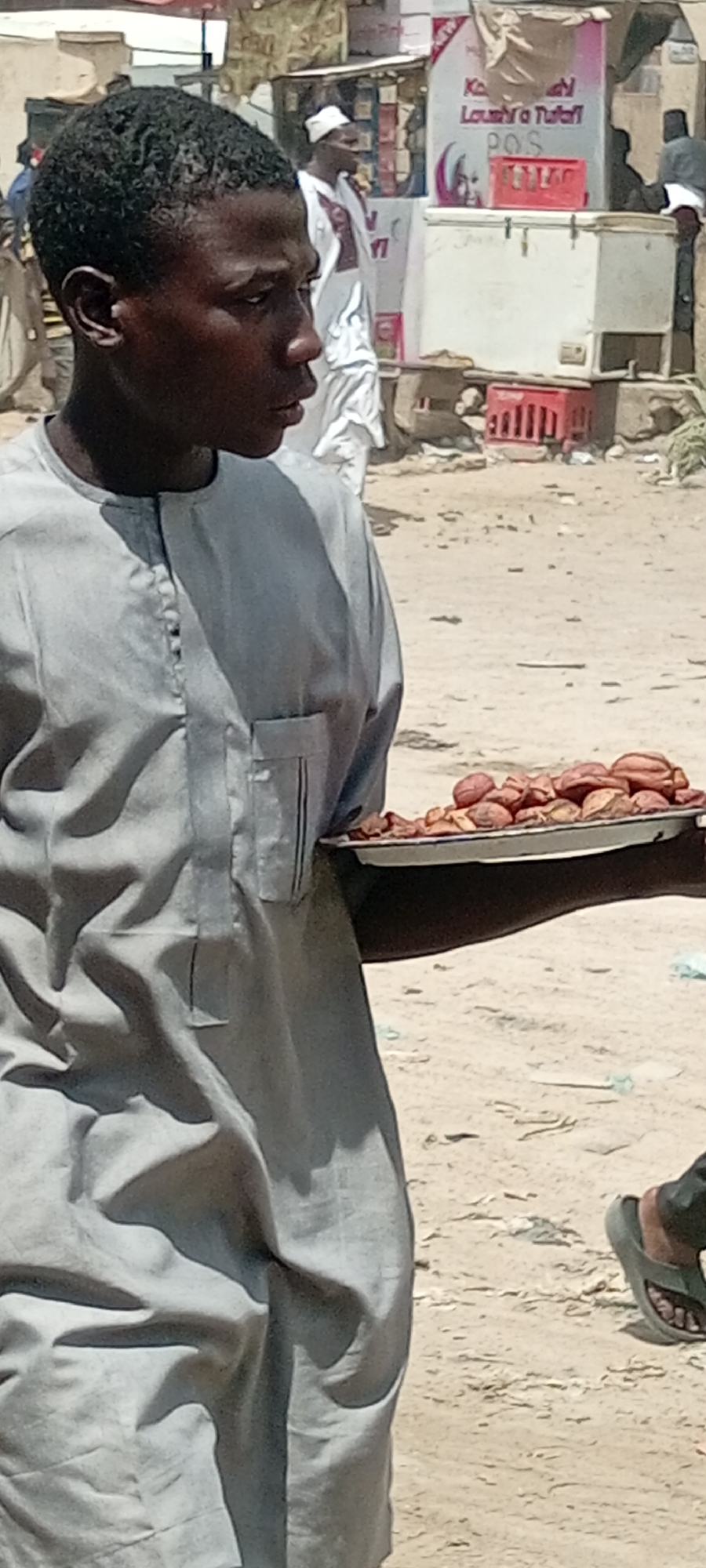In Fagge C, Nigeria, a young man stands on a sandy surface, wearing a light gray traditional shirt. He holds a silver platter filled with red-brown objects, possibly nuts or dates. In the background are other people, one in a white robe and cap, and a small shop with signs in a local language. Red plastic crates are also visible behind the man.
