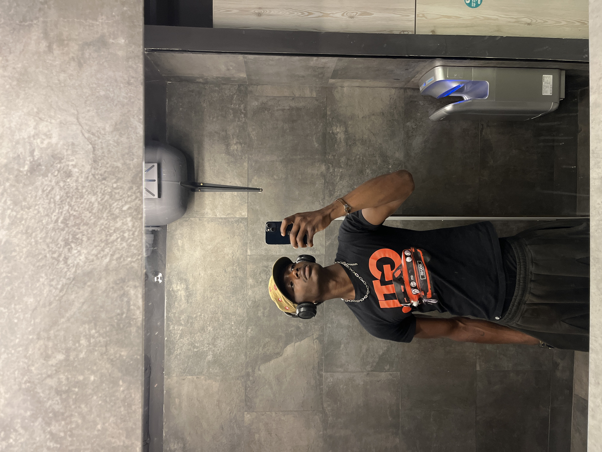 A man is taking a selfie in a bathroom. The man is wearing a black t-shirt, dark sweatpants, a camouflage baseball cap, and black headphones. He also has a silver chain and a bracelet. The bathroom has gray tile walls and floors. A hand dryer is mounted on the wall behind the man. A sink with a faucet is visible to the left. It is unknown whether this photograph was taken in the city of Barcelona, Spain.
