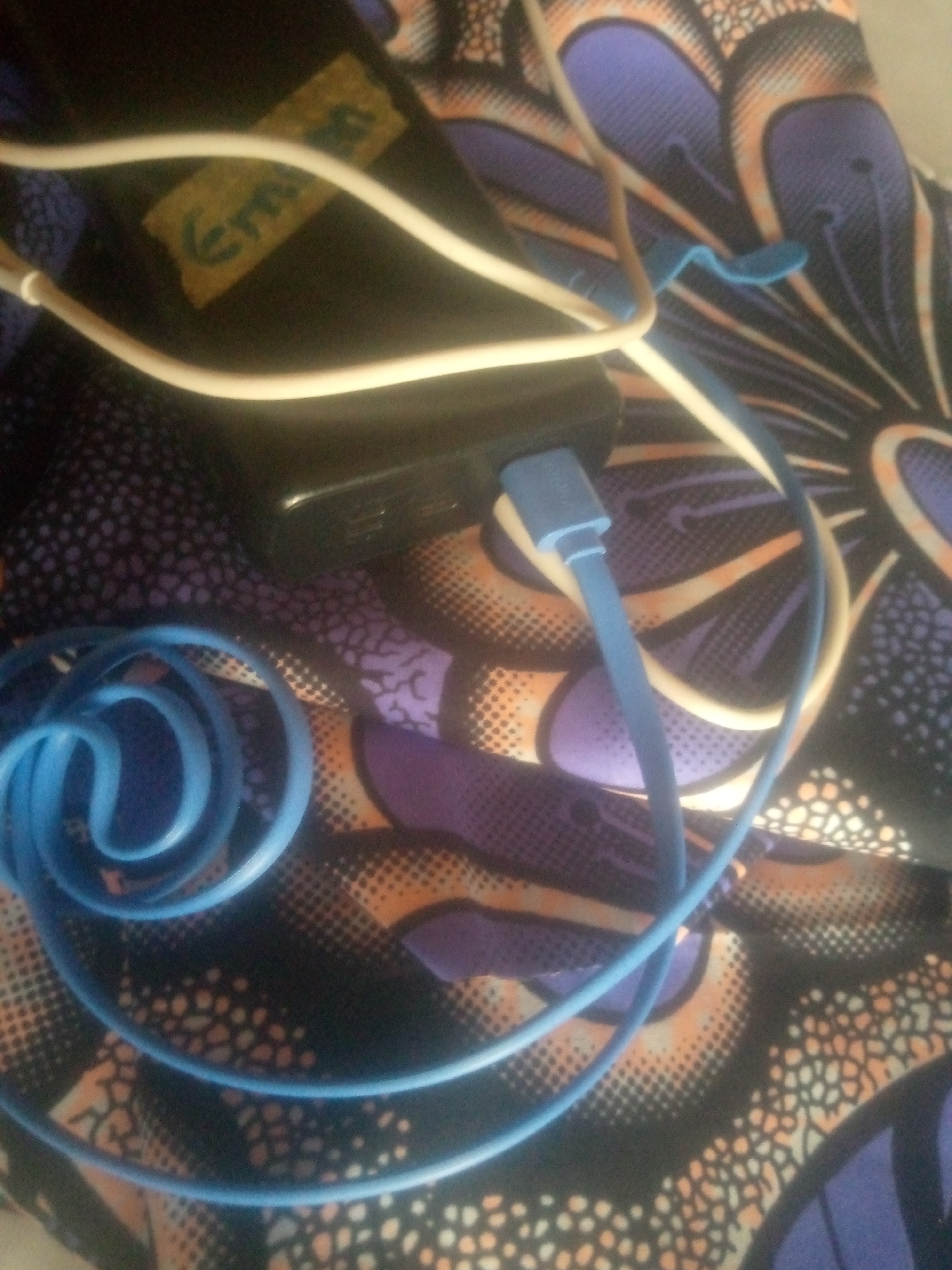 The image shows a close-up view of electronic accessories, specifically a black power adapter and its connected charging cables. One cable is white and appears to be a USB cable. The other cable is bright blue and is coiled in the foreground. The power adapter has a small piece of yellow tape stuck to it with some text written in green marker. The text on the tape appears to read "Emirl". The accessories are placed on a patterned fabric that features large, stylized floral or feather-like motifs in shades of purple, orange, and black. The fabric has a textured appearance, with small dots in some areas. The lighting suggests it is indoors, likely in a domestic setting. No people are visible in the image. The scene is static, with no apparent activity taking place other than the presence of the charging equipment. The image is framed to focus on the texture and colors of the fabric and the details of the cables and adapter.