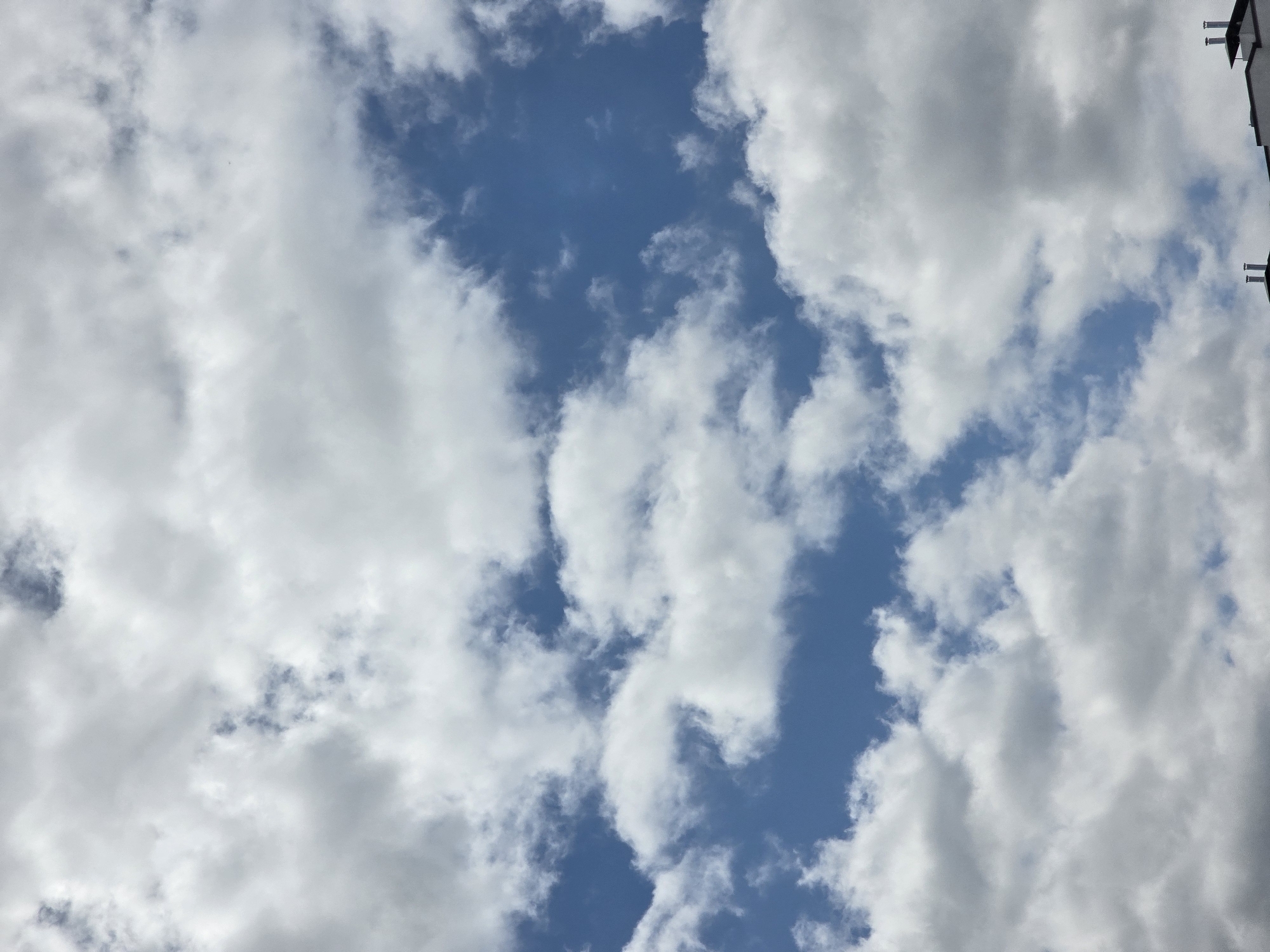 The image is a vertical shot of a partly cloudy blue sky, with several fluffy white clouds scattered across the frame. At the very bottom of the image, the top edge of a building with chimneys is visible. The scene appears to be an outdoor view, likely taken from a residential area. The clouds suggest a fair weather day with some cloud cover. No people or specific actions are depicted.